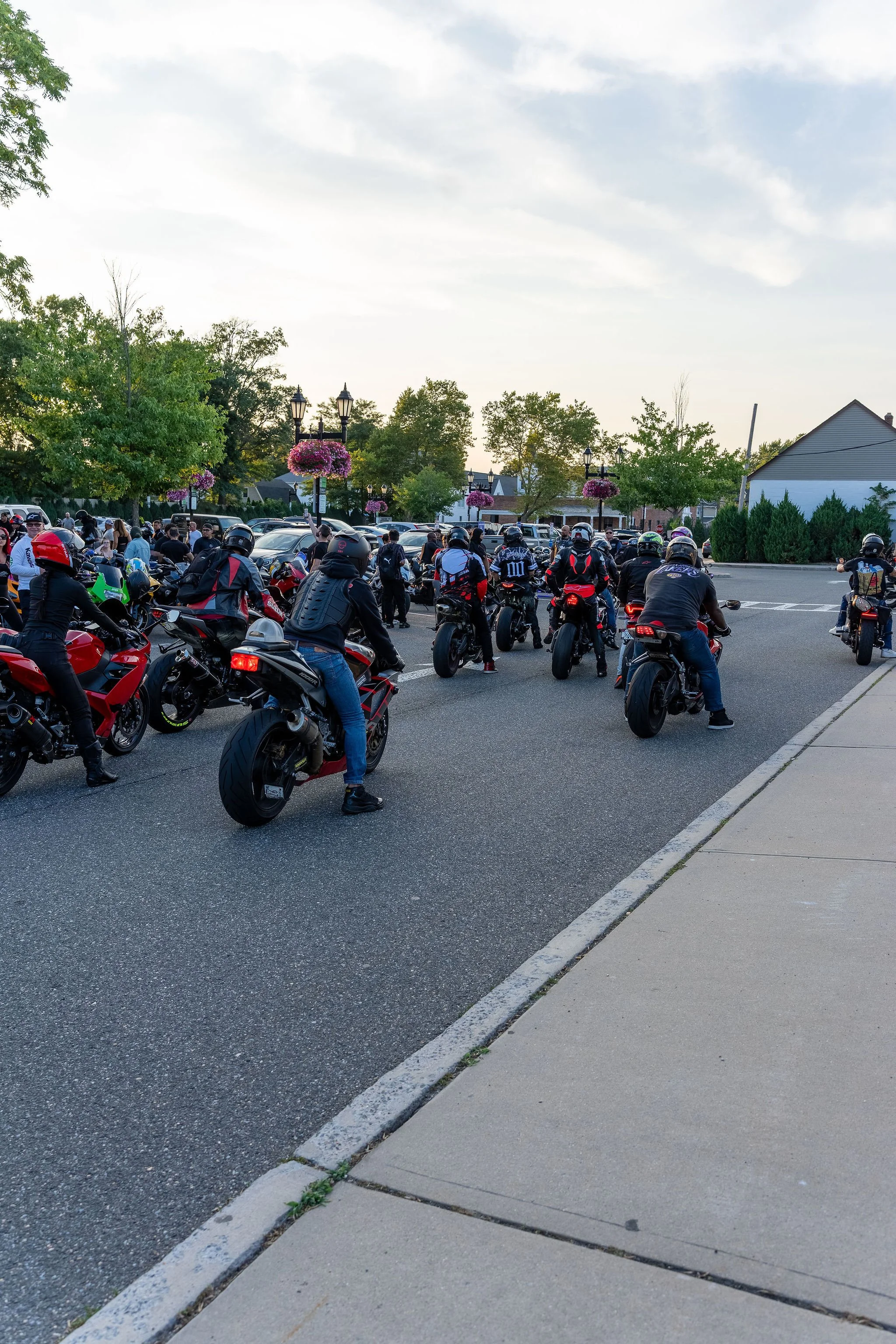 A large group of motorcyclists gathered on a street, with some traffic and residential buildings in the background, under a partly cloudy sky.