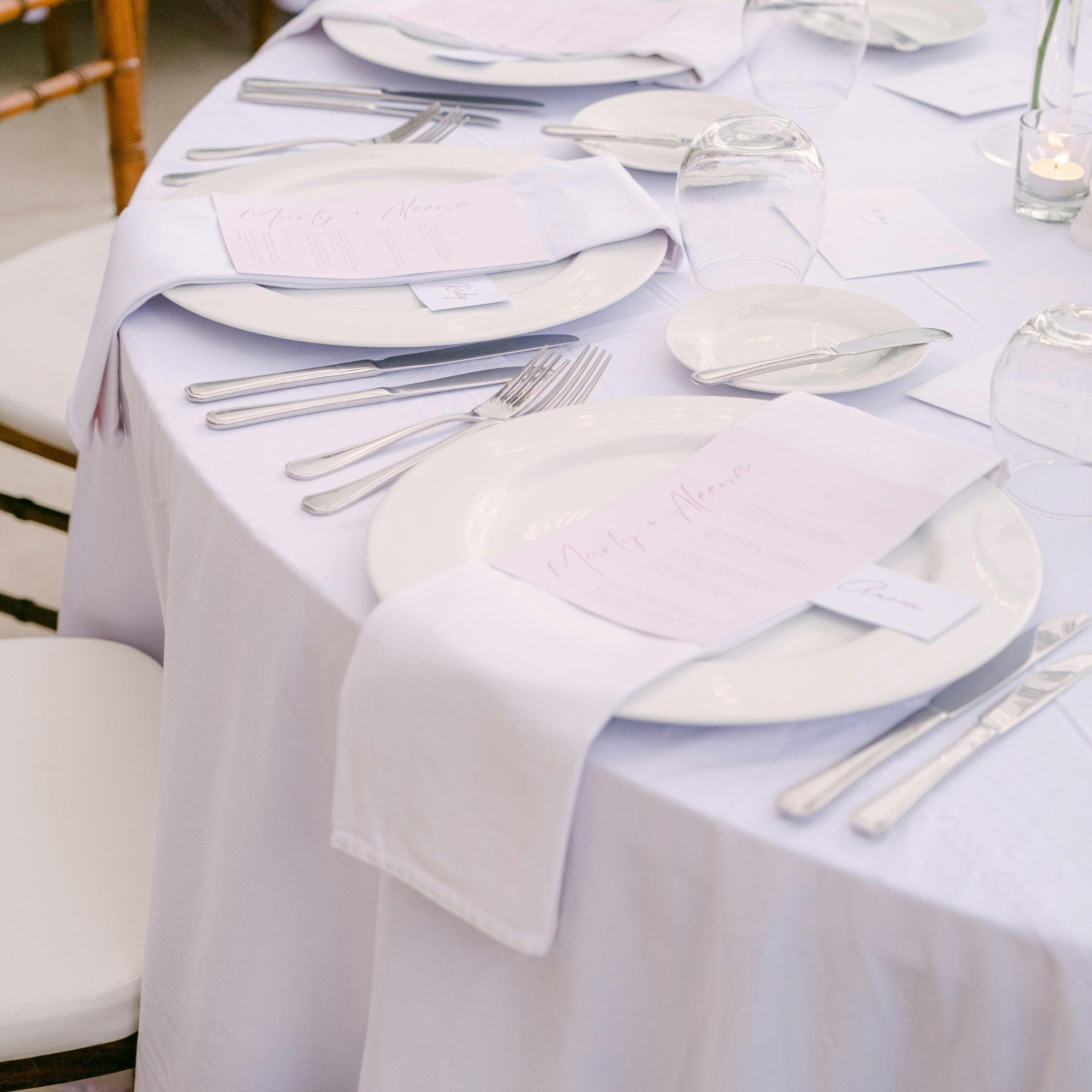 A neatly set dining table with white tablecloth, white plates, silverware, pink place cards, upside-down glasses, and small candles, prepared for an event or celebration.
