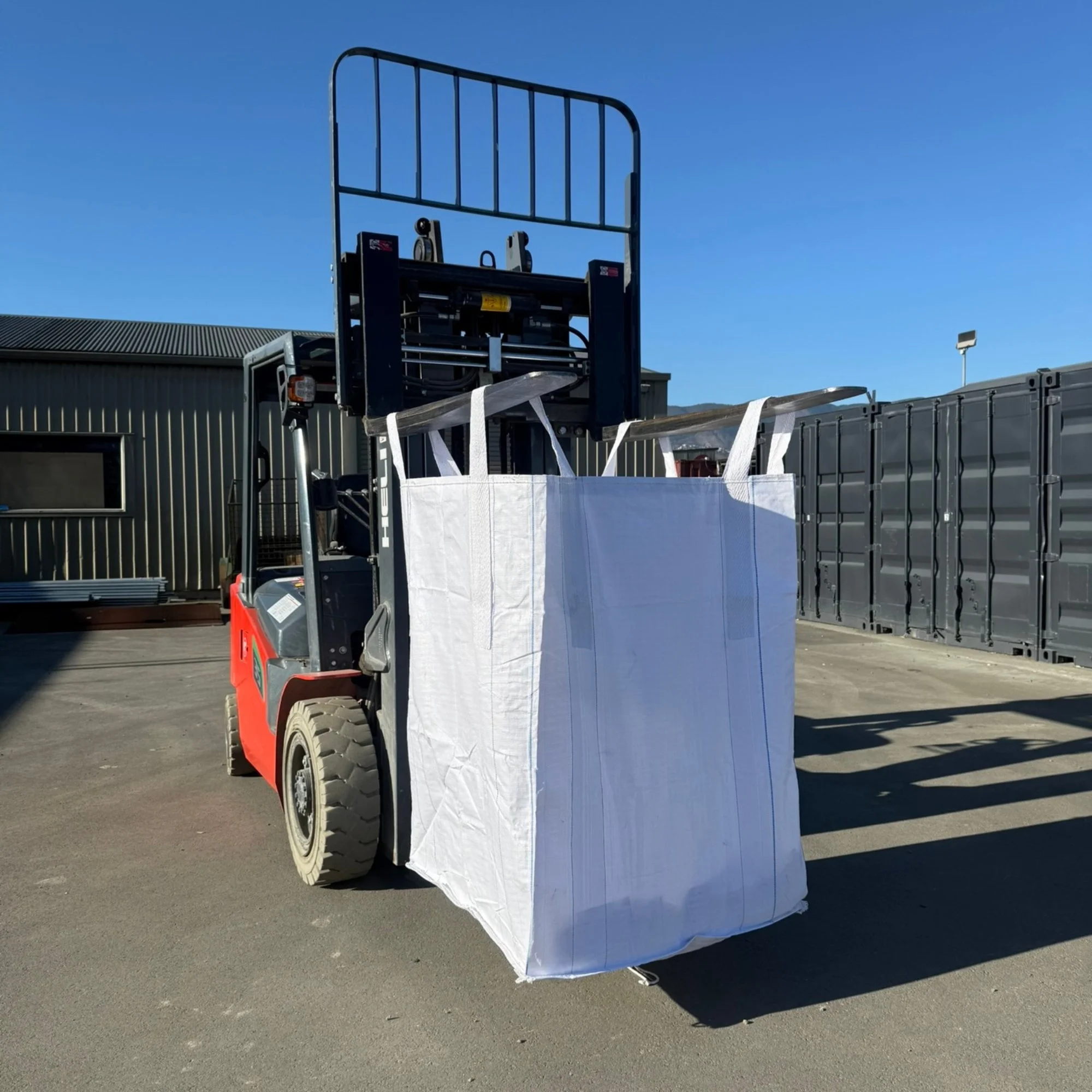 A forklift truck lifting a large white fabric or paper bag outside on a sunny day with a clear blue sky.