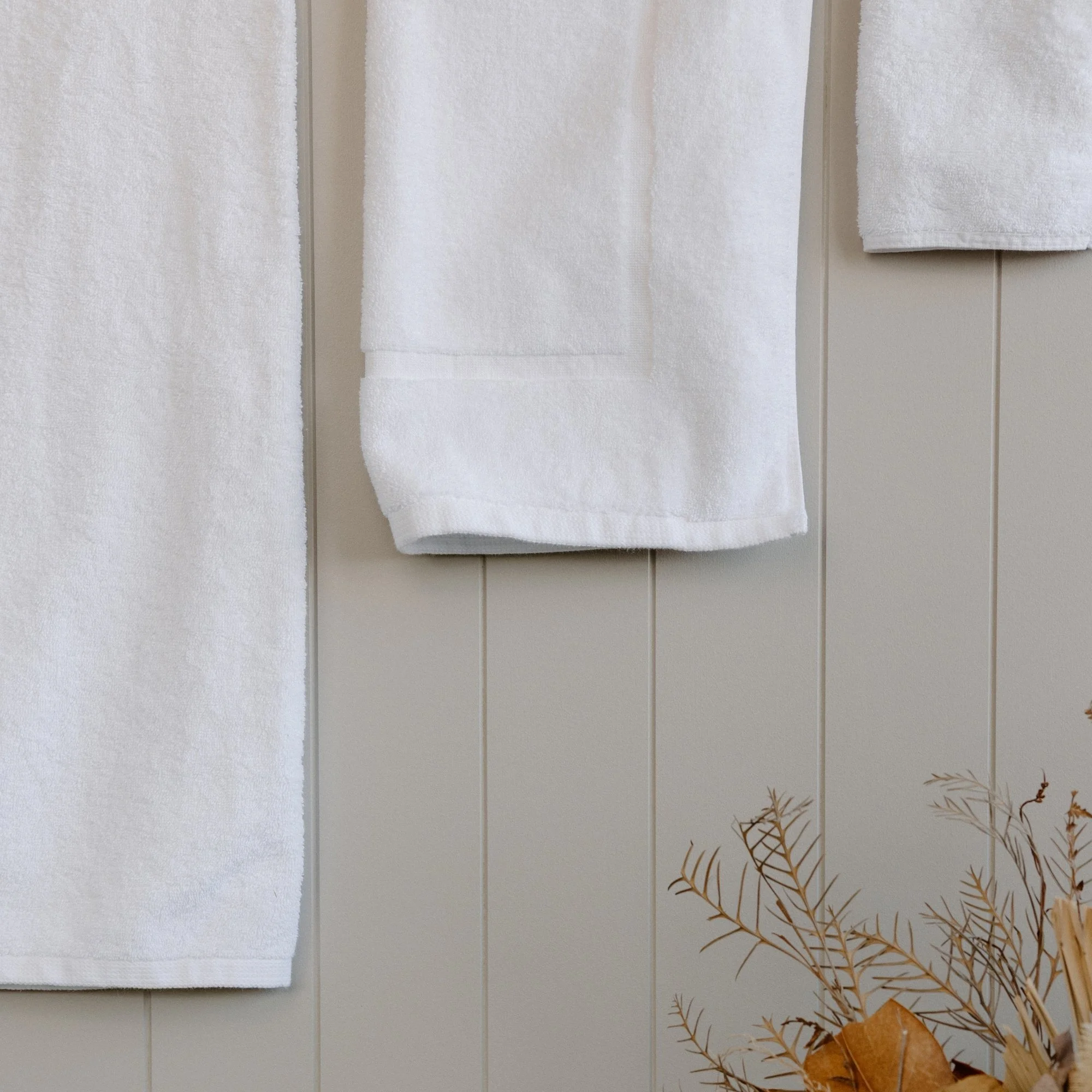 White Tasman bath towels hanging on a beige wall with decorative dried plants in the bottom right corner.