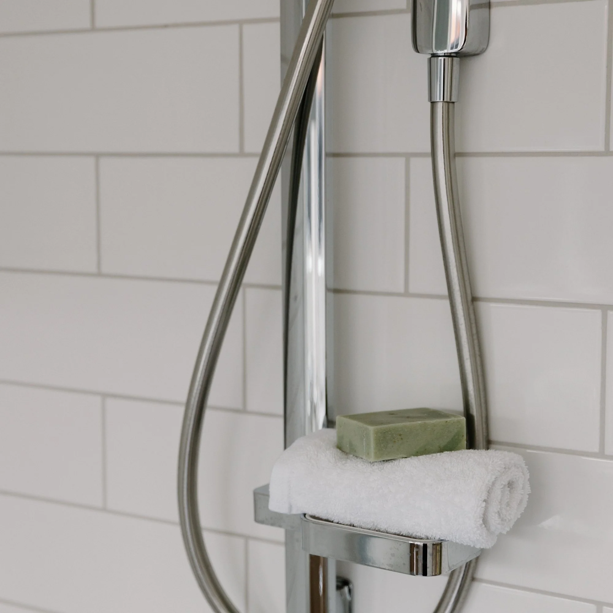 Shower wall with white tiles, a stainless steel shower head, and a soap bar on a rolled white Tasman Facecloth.