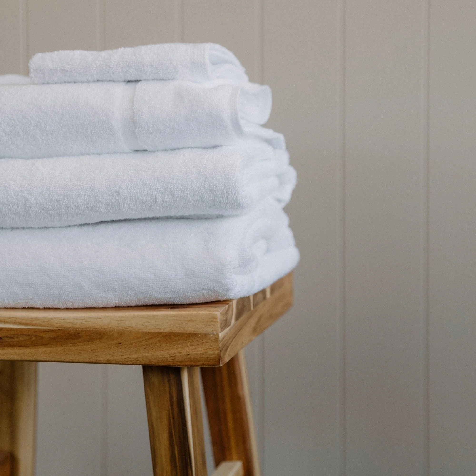 Tasman white towels on a wooden stool in front of a beige paneled wall.