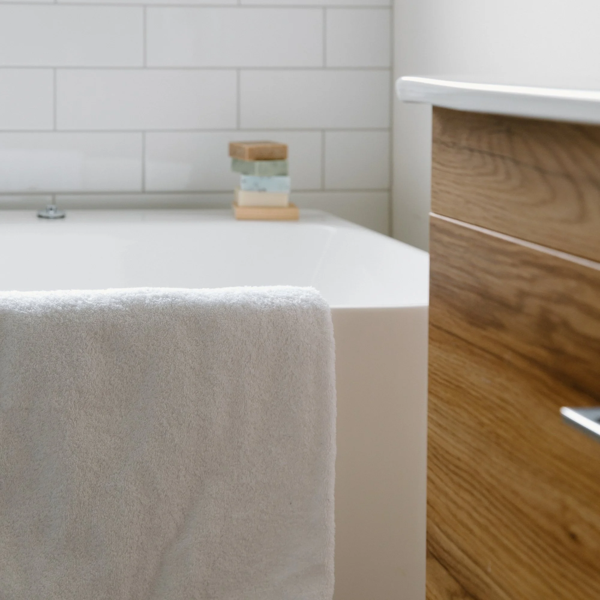 Close-up of a white bathtub with a soft white Tasman bath towel hanging over its edge, a wooden vanity on the right, and a brick-patterned wall in the background with some small stacked stones placed on the edge of the bathtub.