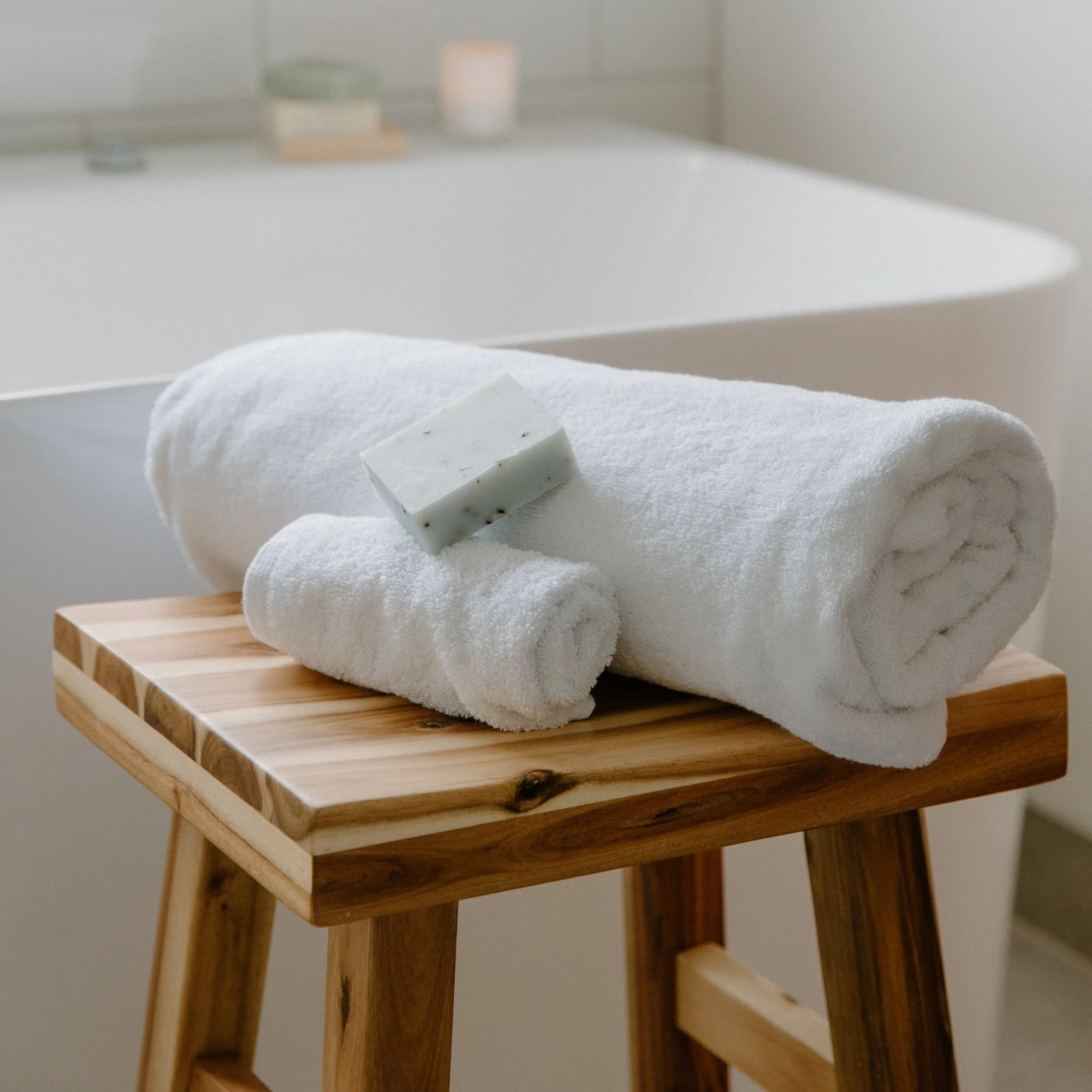 Stacked white Tasman towels with a bar of soap on top, placed on a wooden stool in a bathroom.