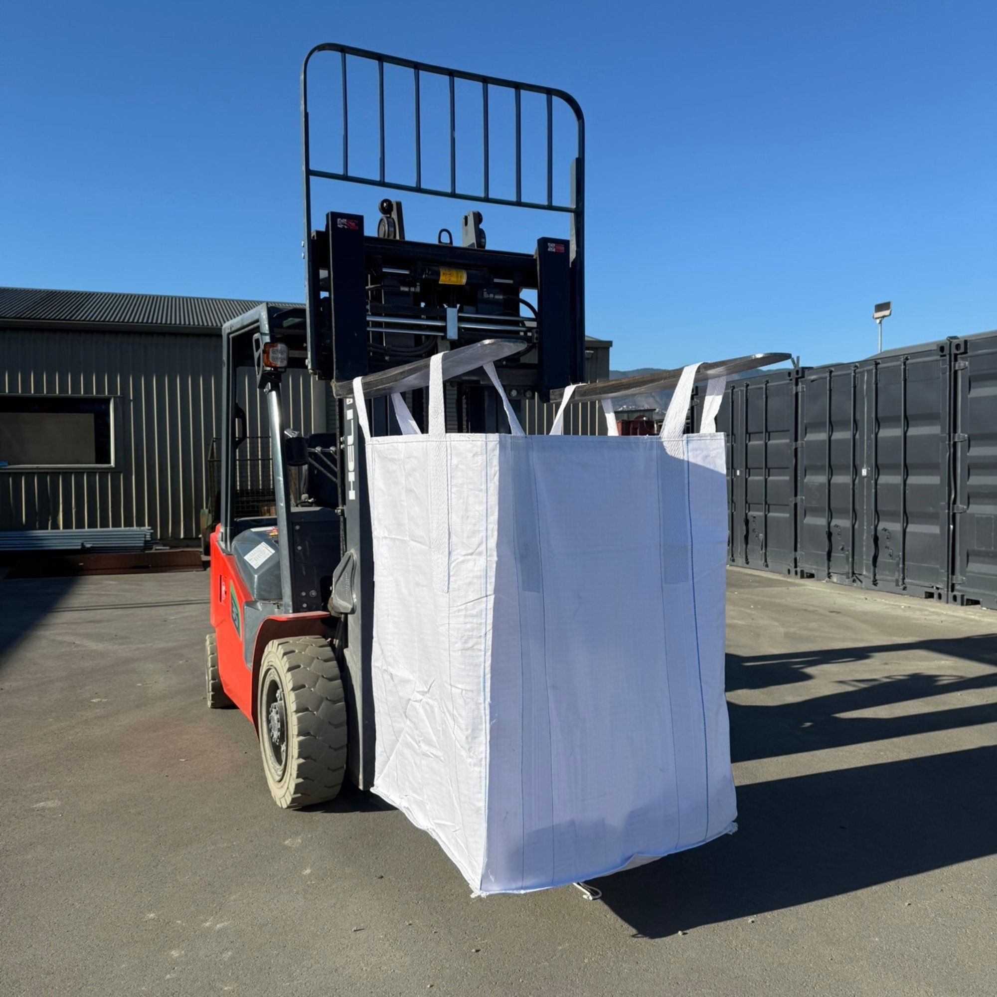 A forklift truck with a white fabric bag attached, situated on an outdoor paved area with a building and containers in the background under a clear blue sky.