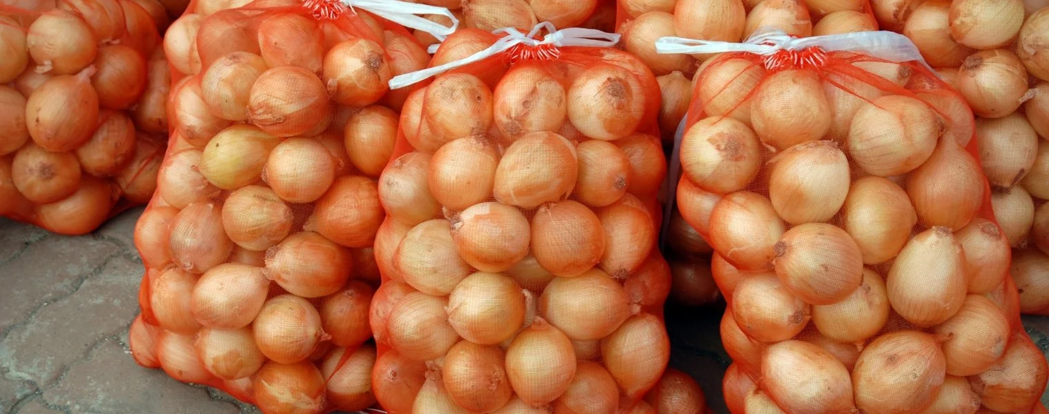 Bags of white onions stacked together on a concrete surface in red woven mesh bags.