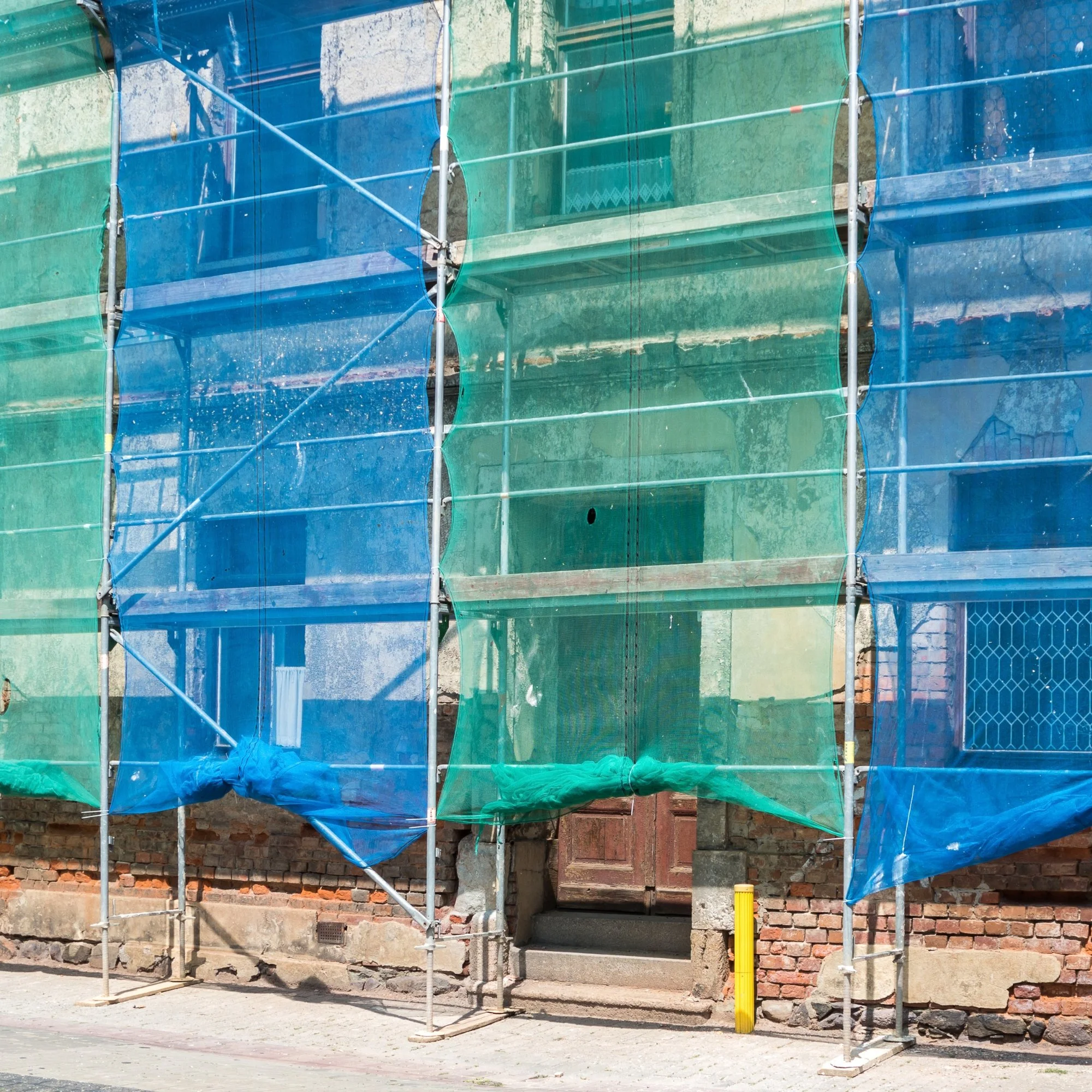 Building under renovation covered with green and blue scaffold netting, with a brick and stone foundation, and a wooden door.