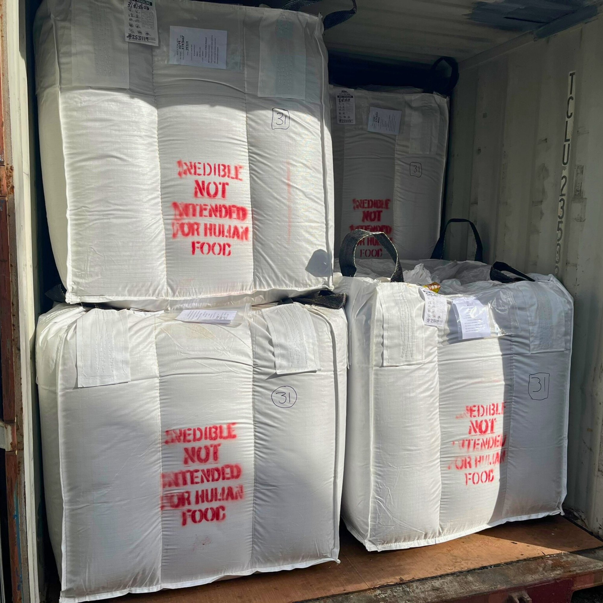 Large white FIBC baffle bags with red spray-painted text reading 'EDIBLE NOT INTENDED FOR HUMAN FOOD', stacked inside a shipping container.