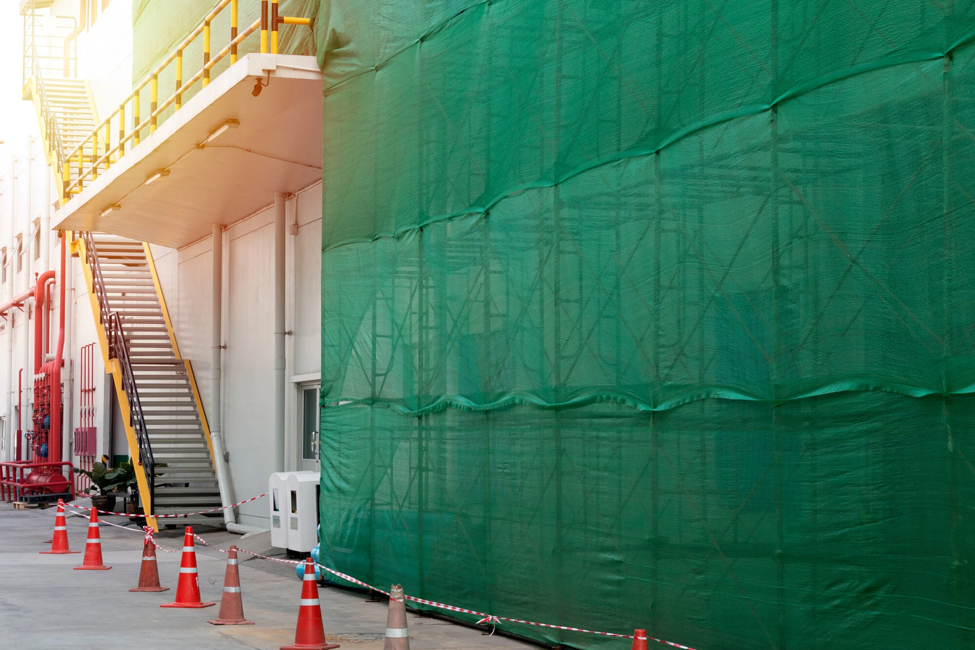 Construction site with green scaffold nettingnetting on scaffolding, orange traffic cones in a row on the ground, a small white window, and exposed red pipes on the left side.