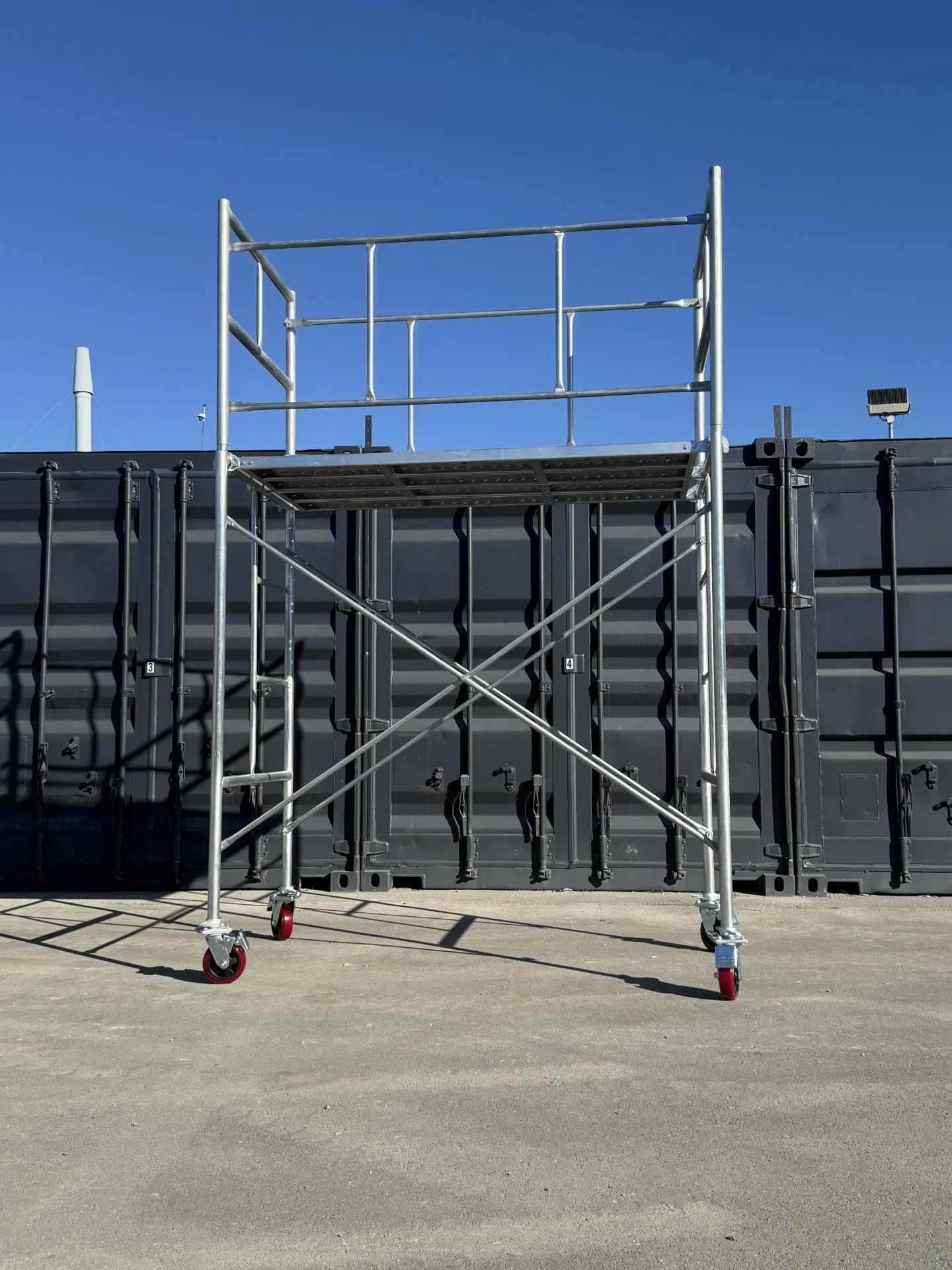 Empty mobile scaffolding tower with four wheels in front of a black wall and a clear blue sky.