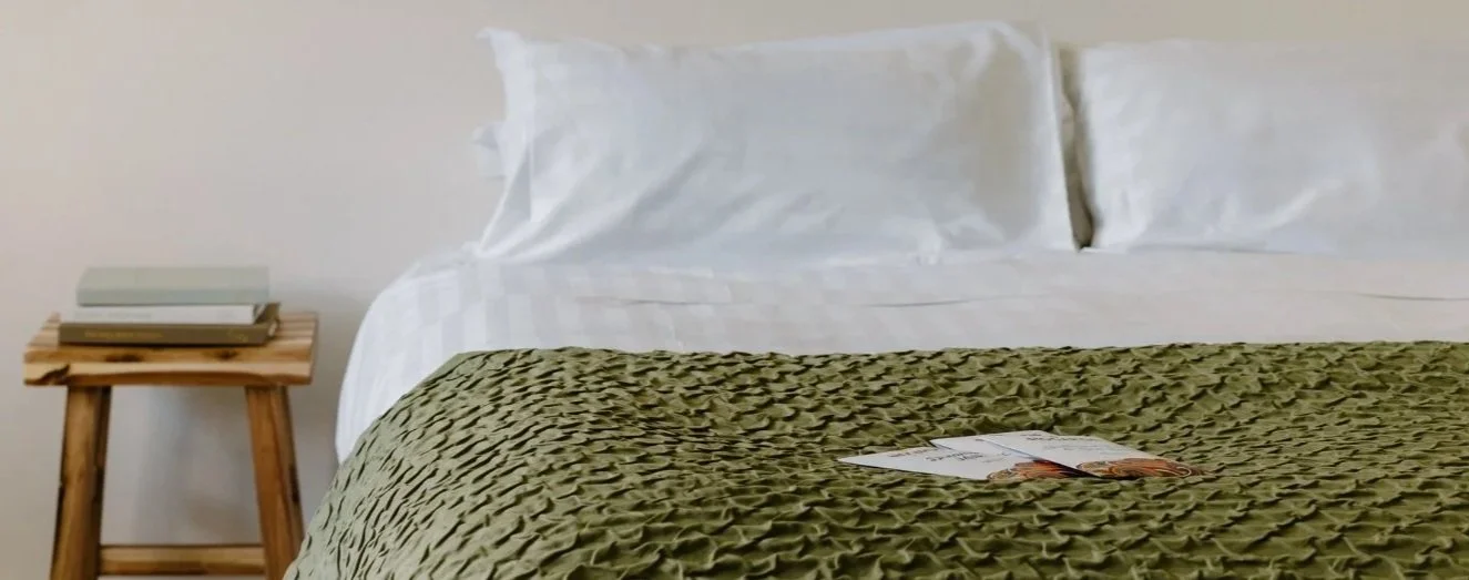A neatly made bed with white pillows and sheets, a green textured blanket, and an open magazine on top, next to a small wooden nightstand with a few books on it.