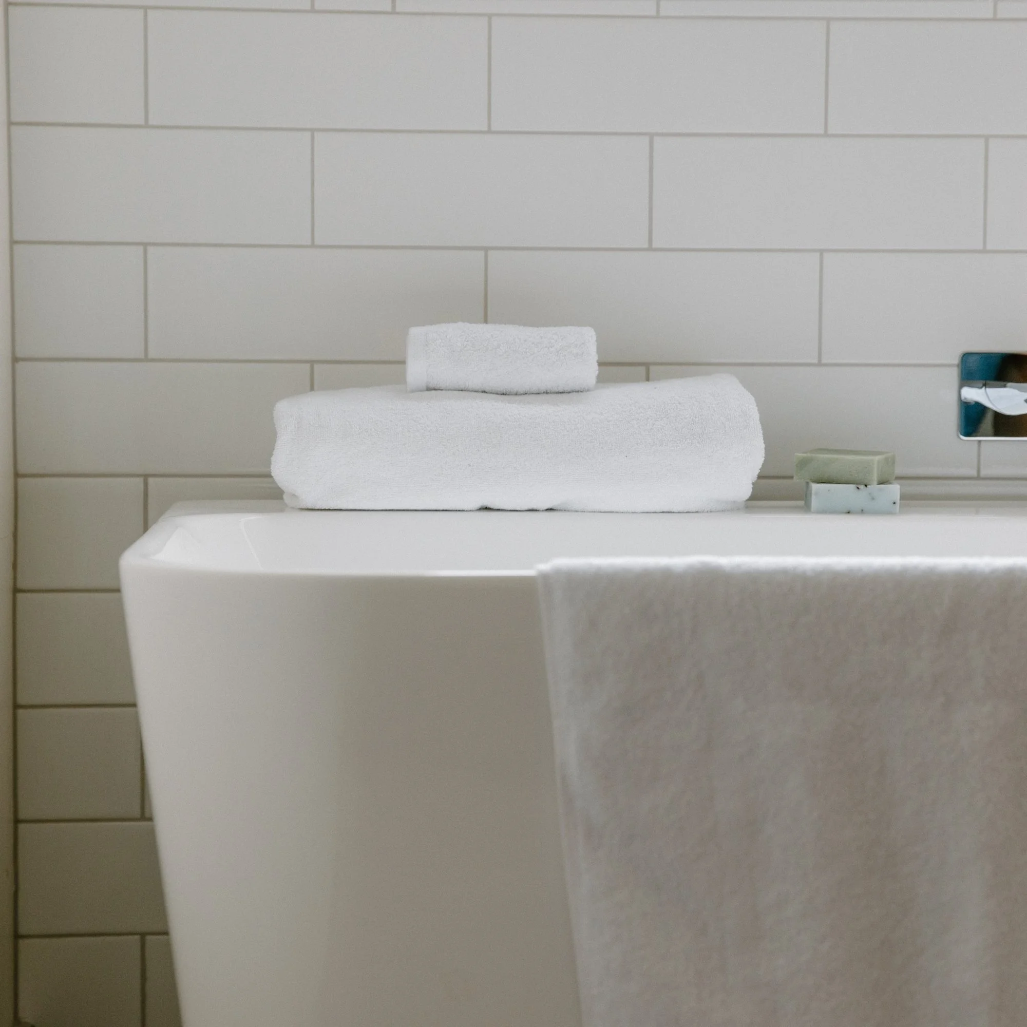 Stacked Tasman white towels on a bathroom sink with tiled wall background.