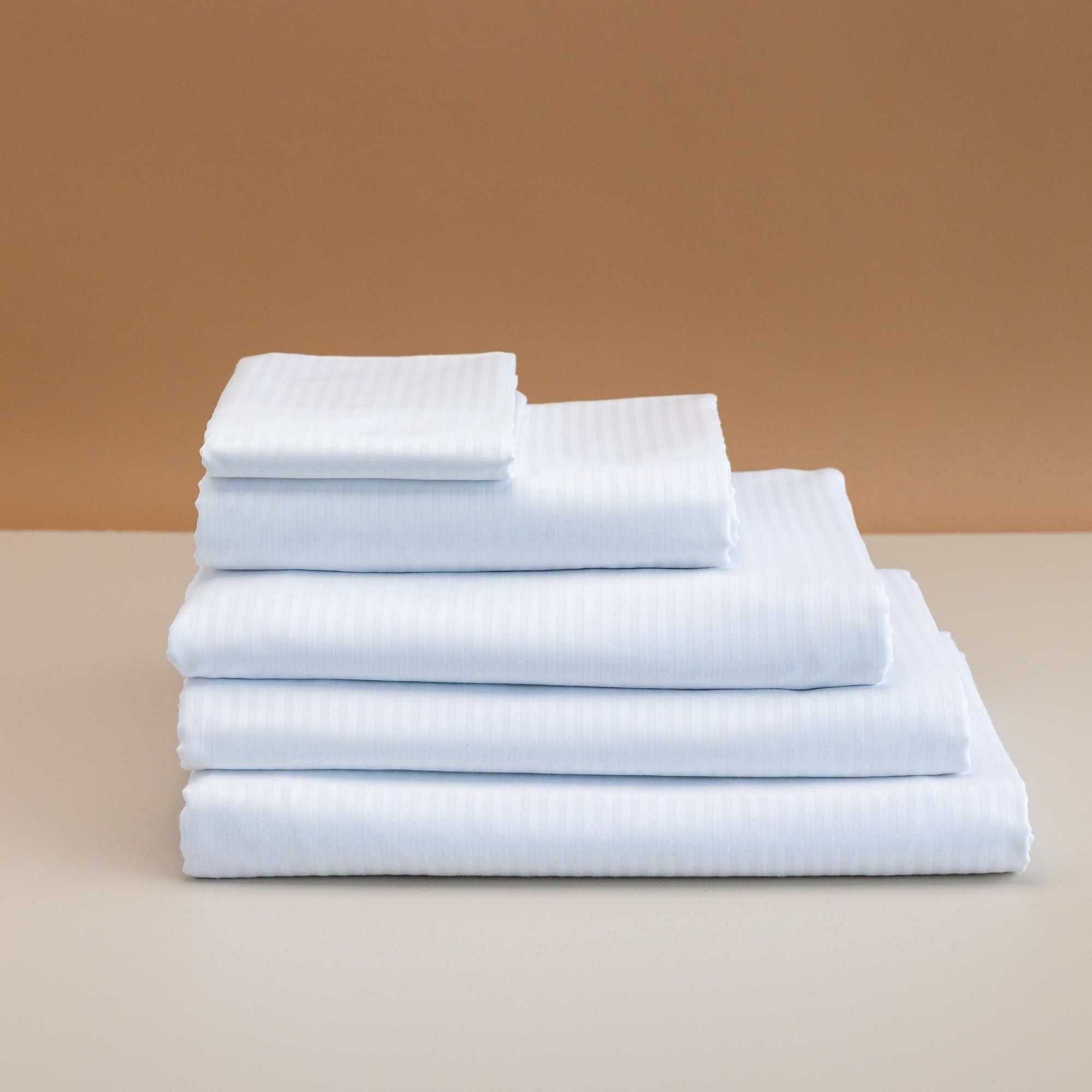 Stack of white Heritage Cotton Rich bed linens on a light-colored surface with a brown and beige background.