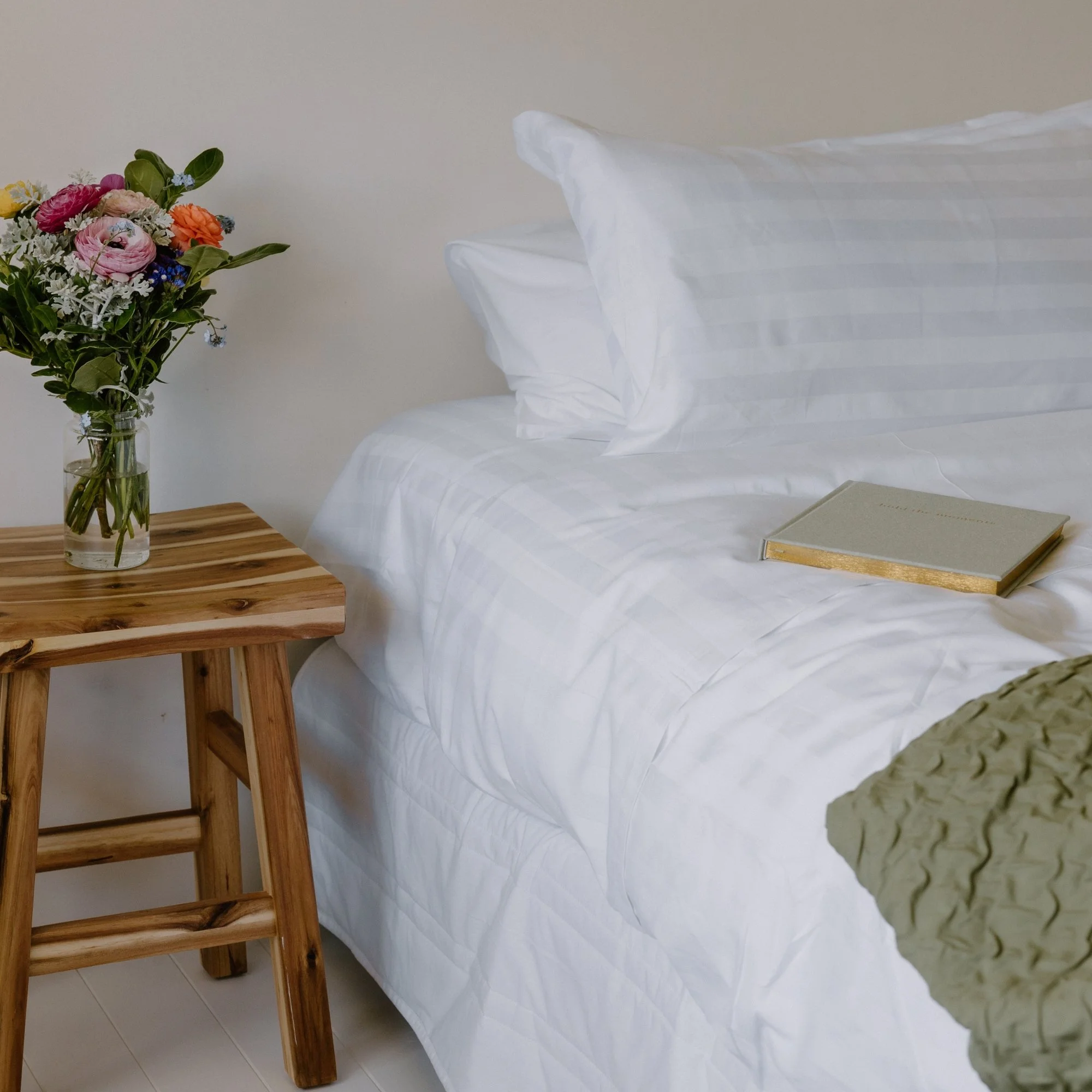 A cozy bedroom with a bed made using Rutherford 25mm Striped sheets, wooden side table, and a glass vase of colorful flowers.