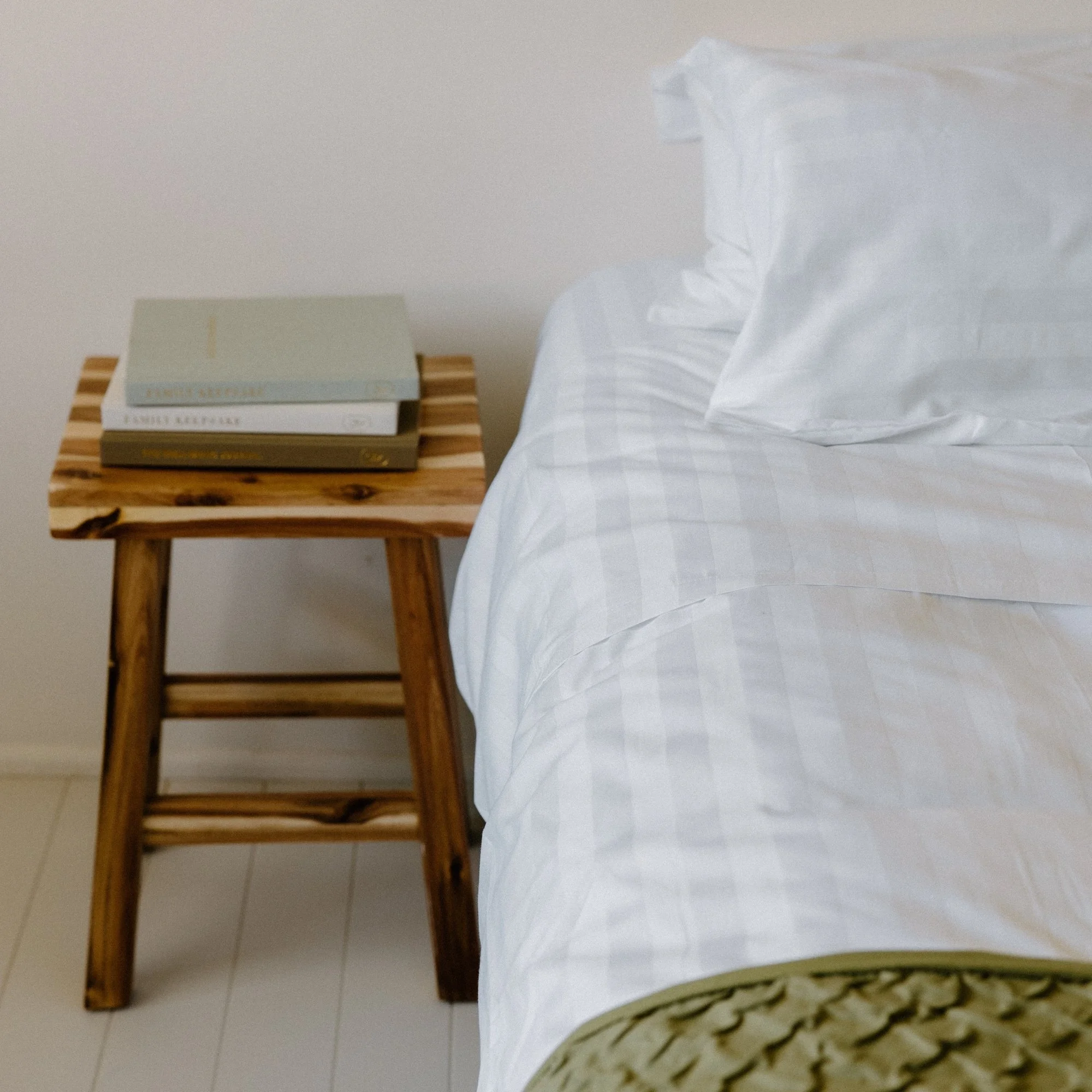 A wooden bedside table with three stacked books next to a bed with white 25mm Rutherford Striped Sheet set.
