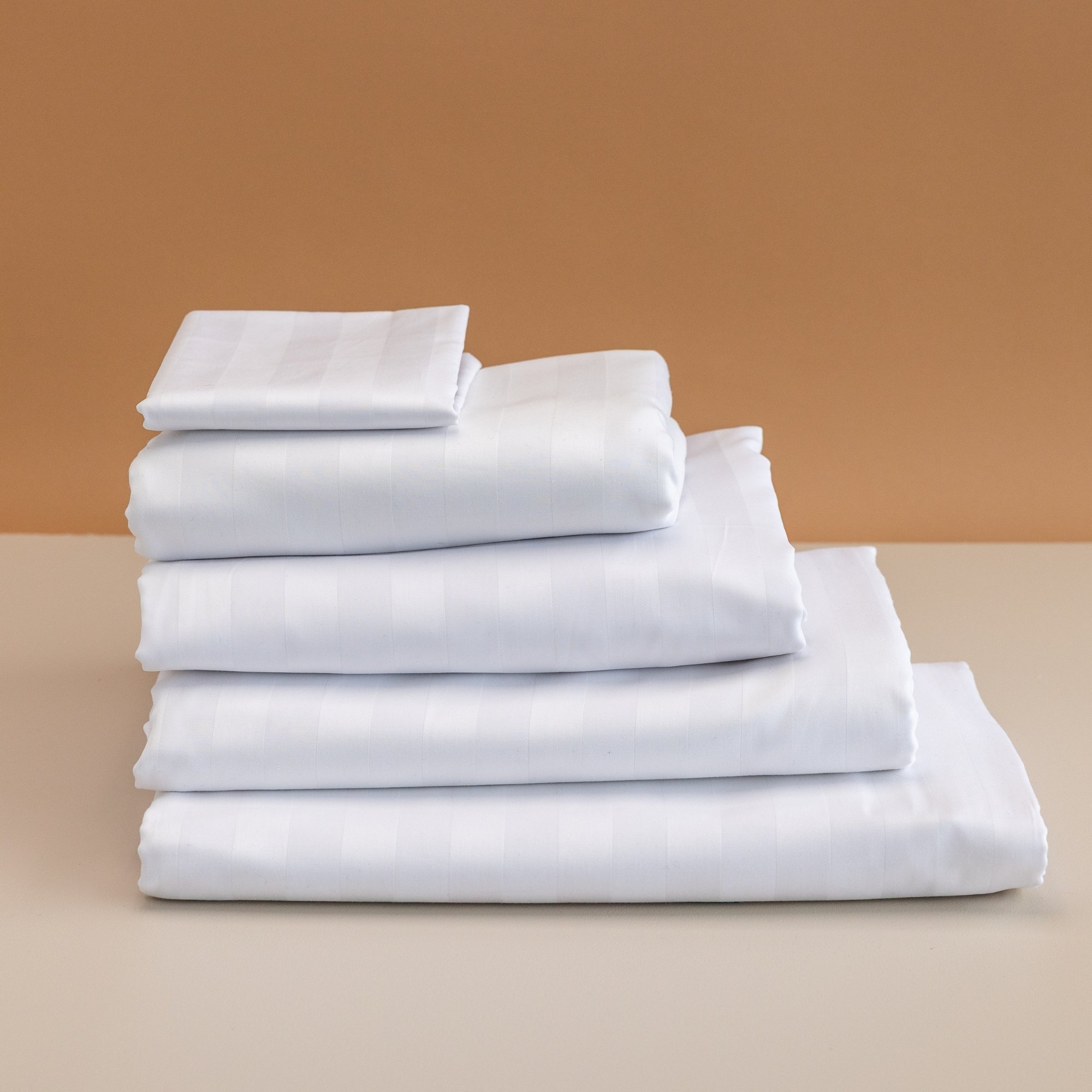 Stack of neatly folded white 25mm Rutherford Striped Sheet sets on a beige surface with a two-tone beige background.
