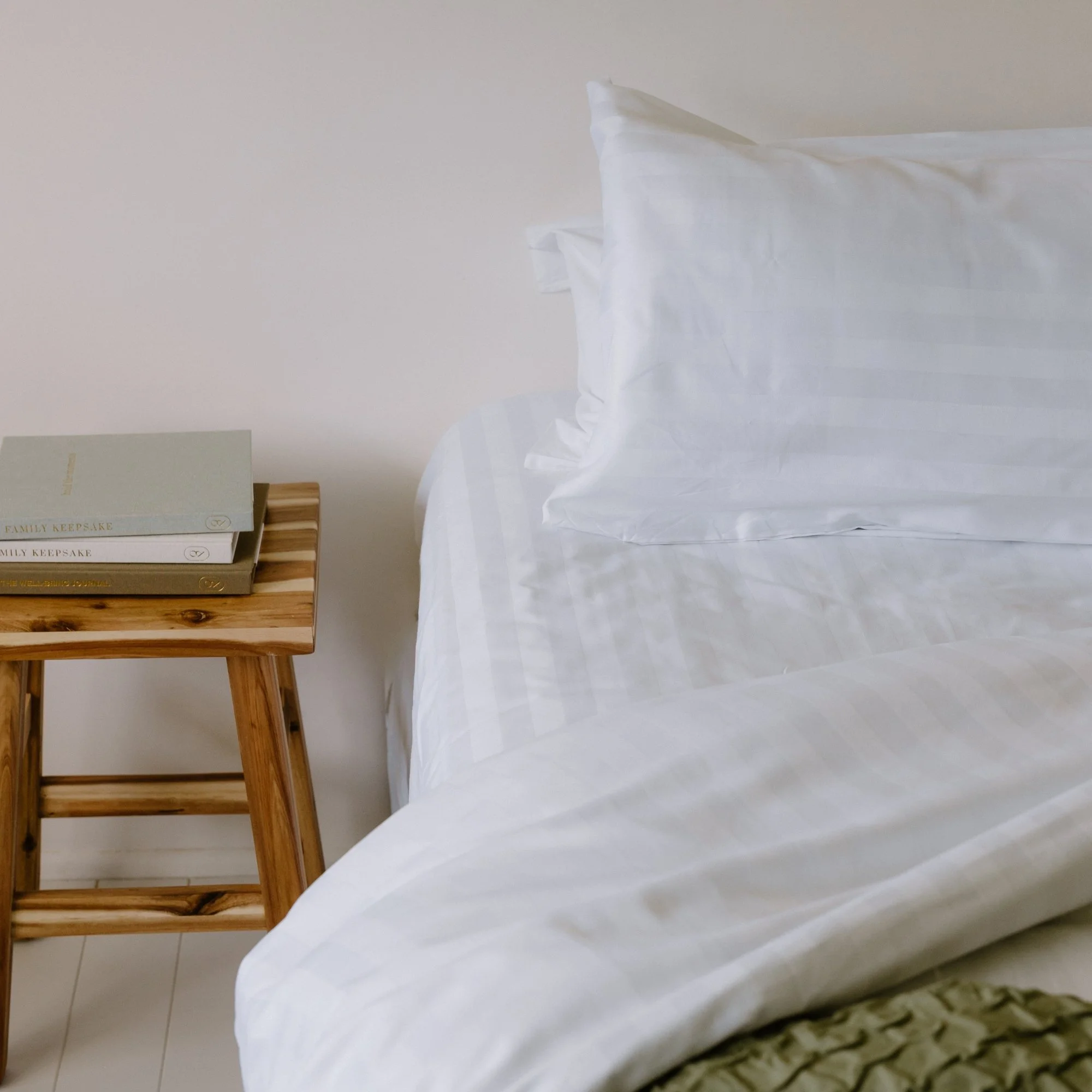 A neatly made bed with white striped sheets and pillows, next to a wooden nightstand with stacked books on top.