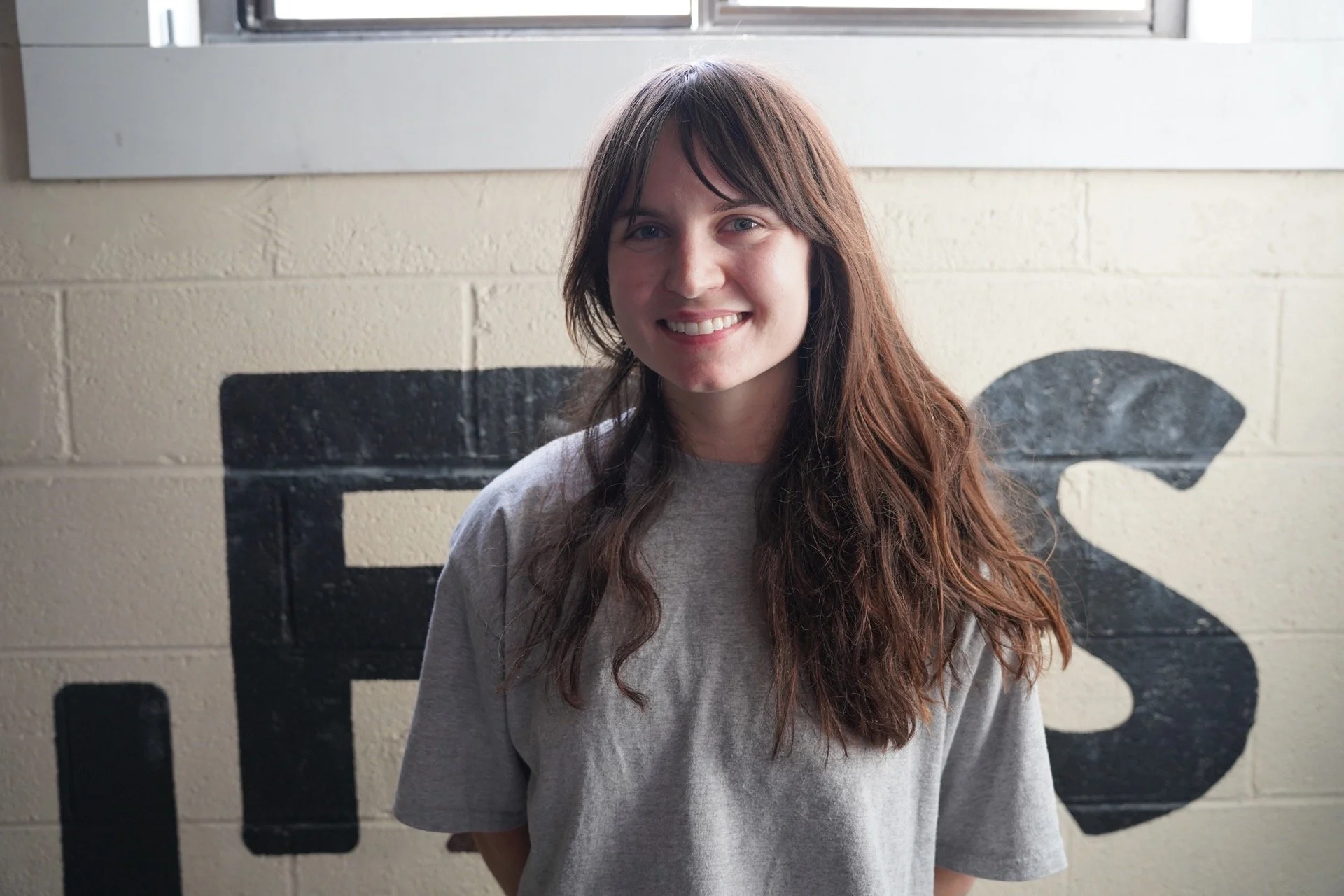 A young woman with long wavy brown hair and blue eyes smiling, standing in front of a wall with black and white lettering visible behind her.