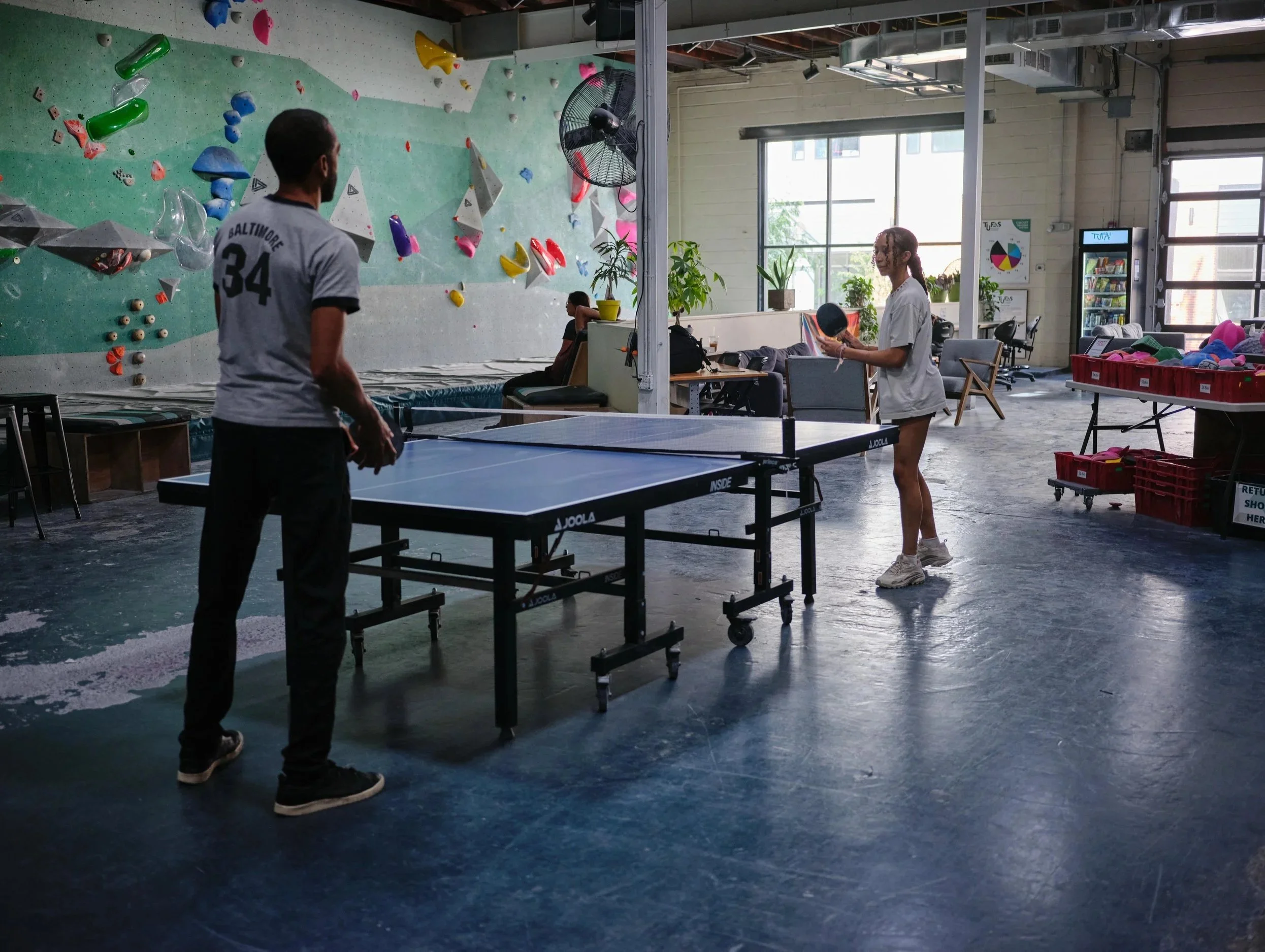 A person standing by a ping pong table in an indoor recreational space. In the background, there is a rock climbing wall with various colorful holds and shapes. Two women are sitting at a table, with one of them reading a magazine. The space is well-
