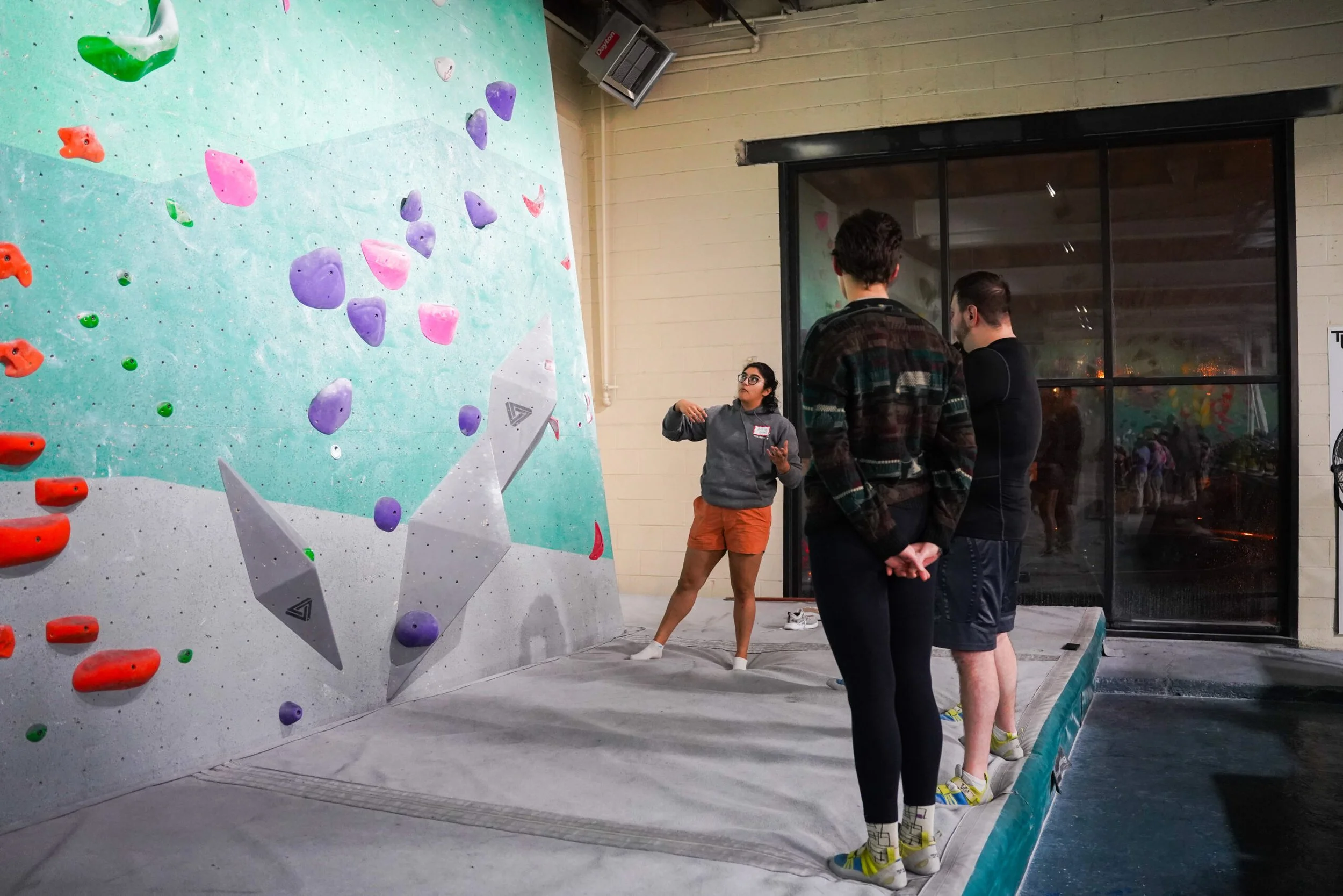 An indoor rock climbing gym with a climbing wall featuring colorful holds. Three people, two men and a woman, are standing and talking near the climbing wall. The woman is gesturing while speaking, and the men are listening. The gym has a large glass window reflecting the climbing area, with a visible safety mat on the floor.
