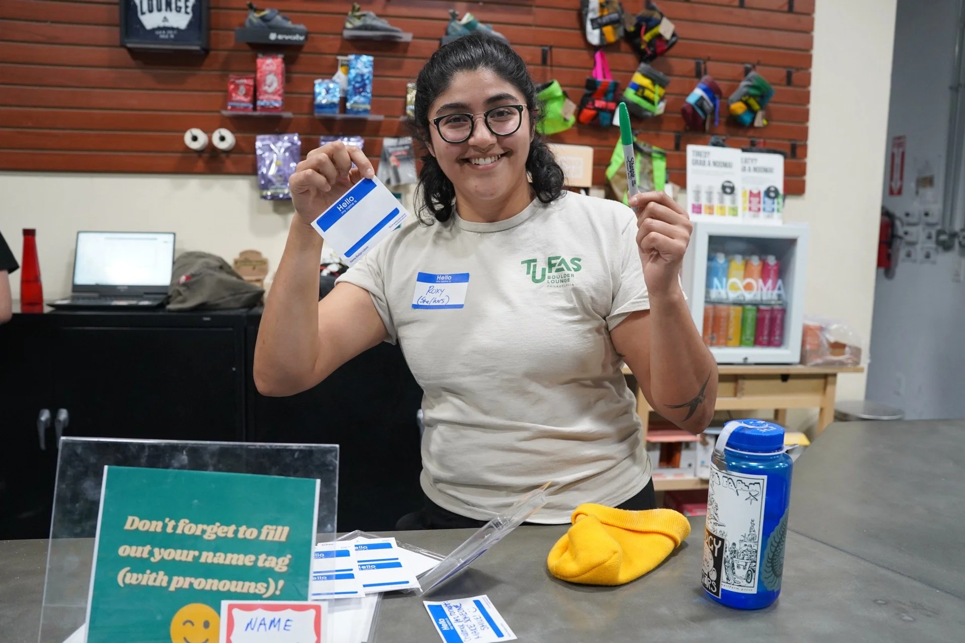 A smiling woman with dark hair and glasses holding a name tag and a green Sharpie marker in a room with shelves of colorful items and shoes in the background. She is wearing a beige T-shirt with a green logo.