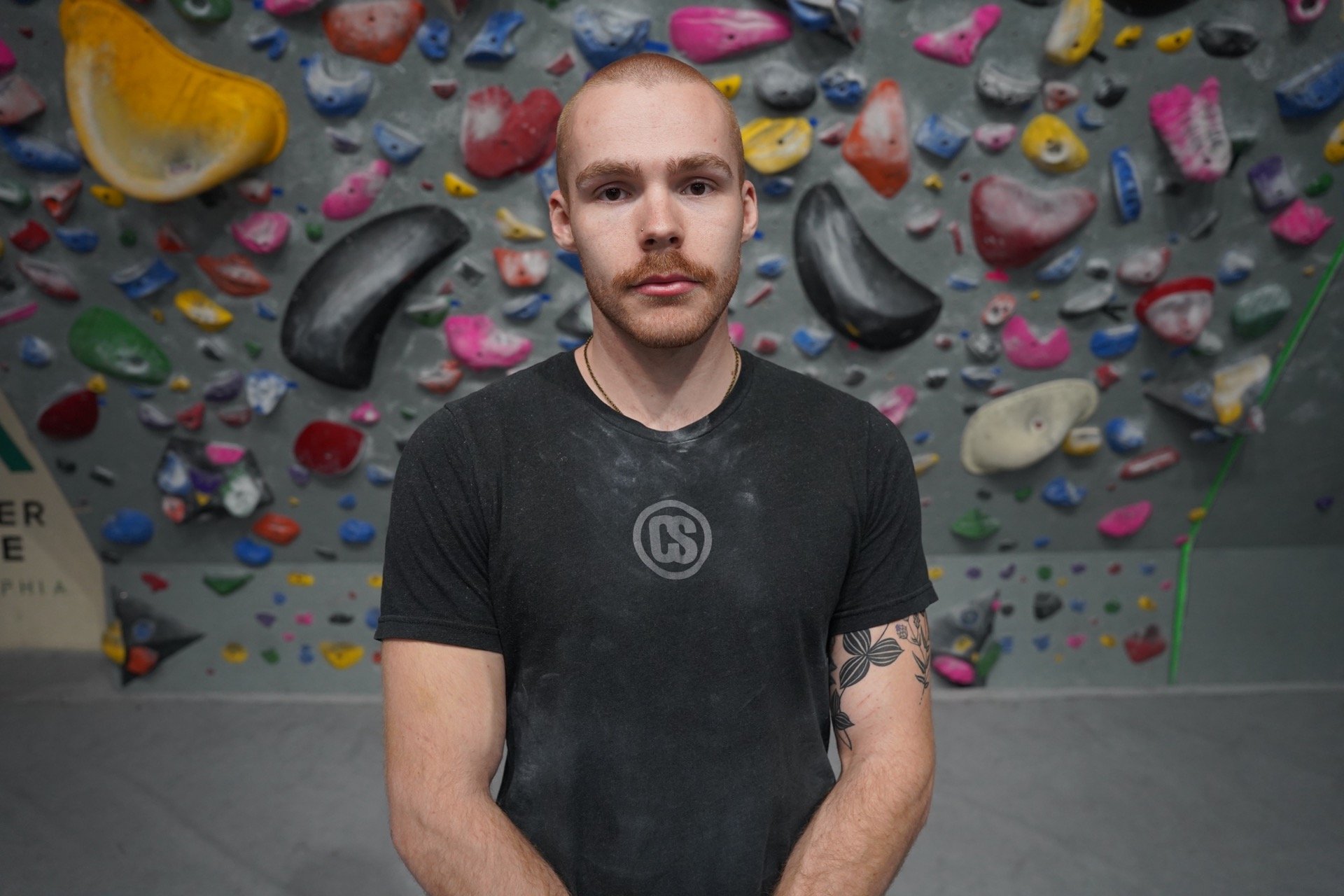 A young man with a shaved head and a beard standing in front of a multicolored indoor climbing wall.