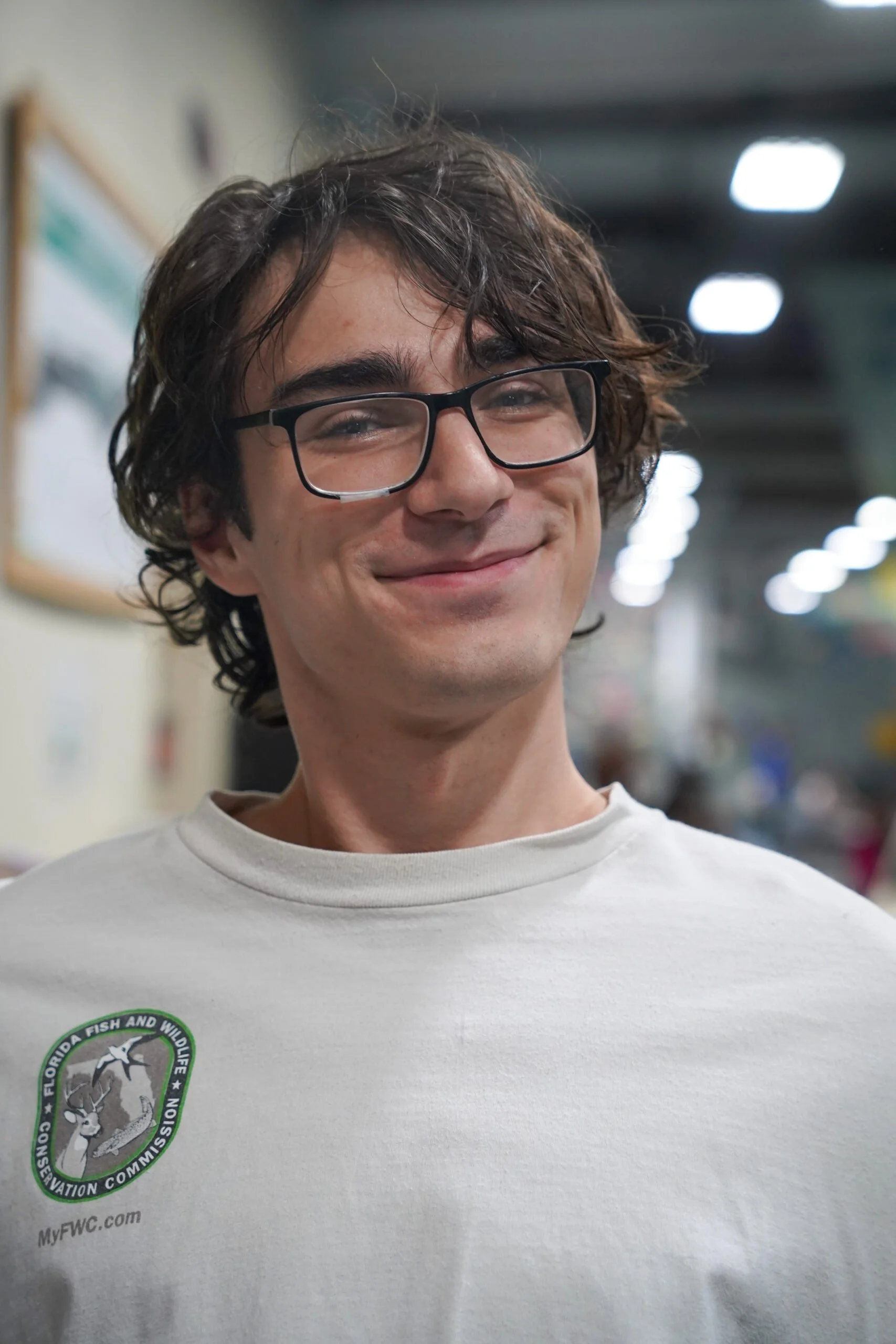 A smiling young man with curly brown hair and glasses, wearing a white T-shirt with a patch that has a logo for the Florida Fish and Wildlife Conservation Commission, standing indoors with bright lights in the background.