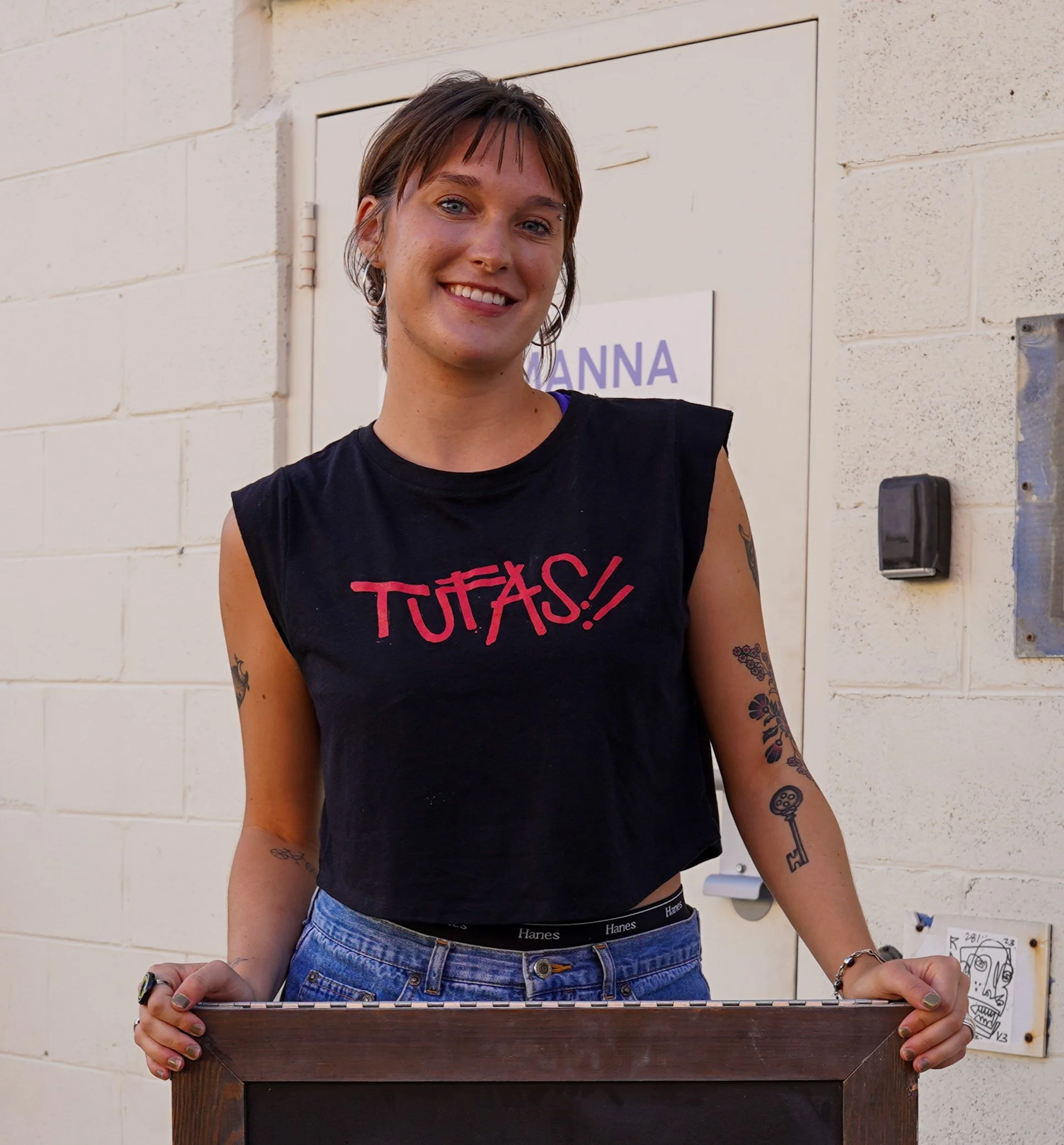 A young woman with short, dark hair and tattoos on her arms, smiling and leaning on a wooden surface, standing outdoors against a beige brick wall with a partially visible sign that has purple text.