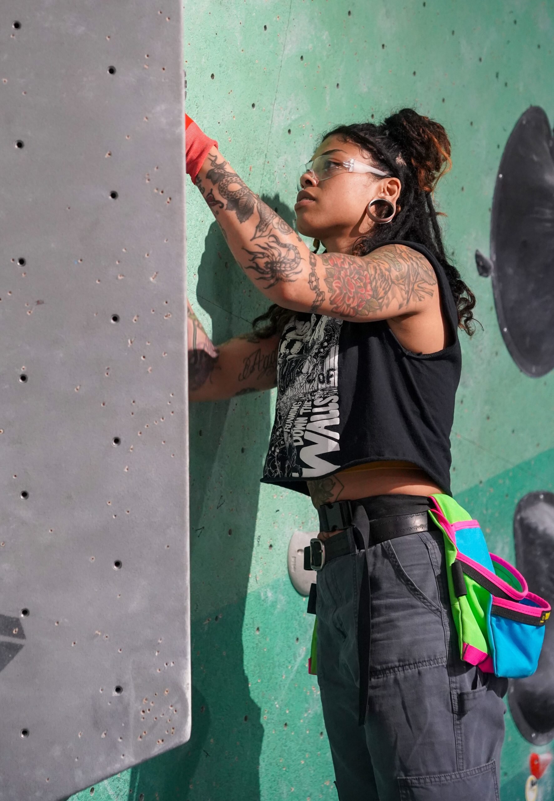A woman with tattoos on her arms and face climbing a grey indoor rock climbing wall with green accents. She is wearing safety glasses, a black graphic t-shirt, and black pants, with a colorful harness or chalk bag around her waist.