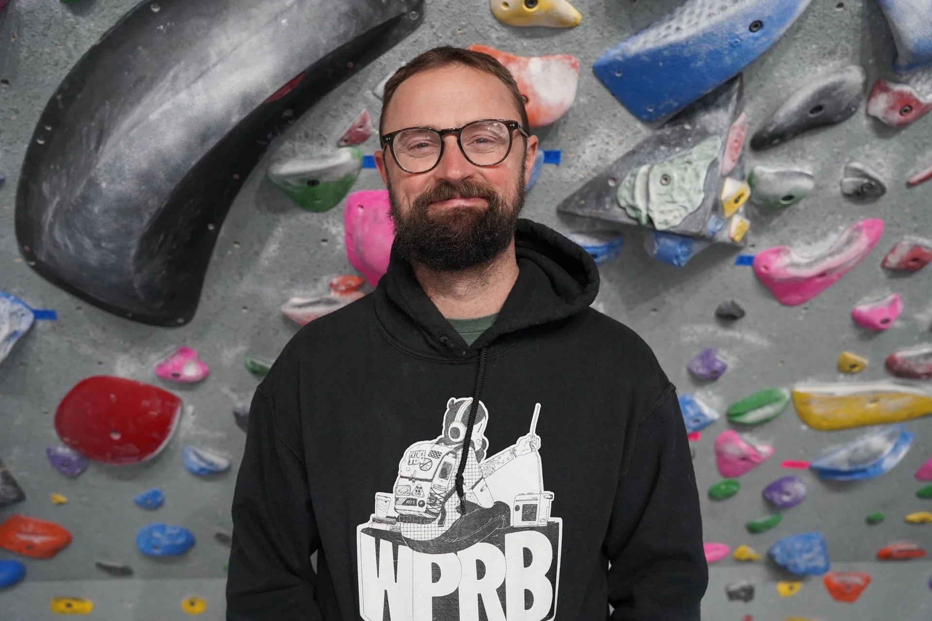 Man with glasses and beard standing in front of a colorful indoor rock climbing wall.