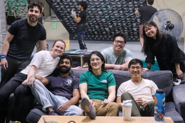 A group of seven young adults, five men and two women, sitting and standing around a gray sofa in an indoor climbing gym, smiling at the camera.