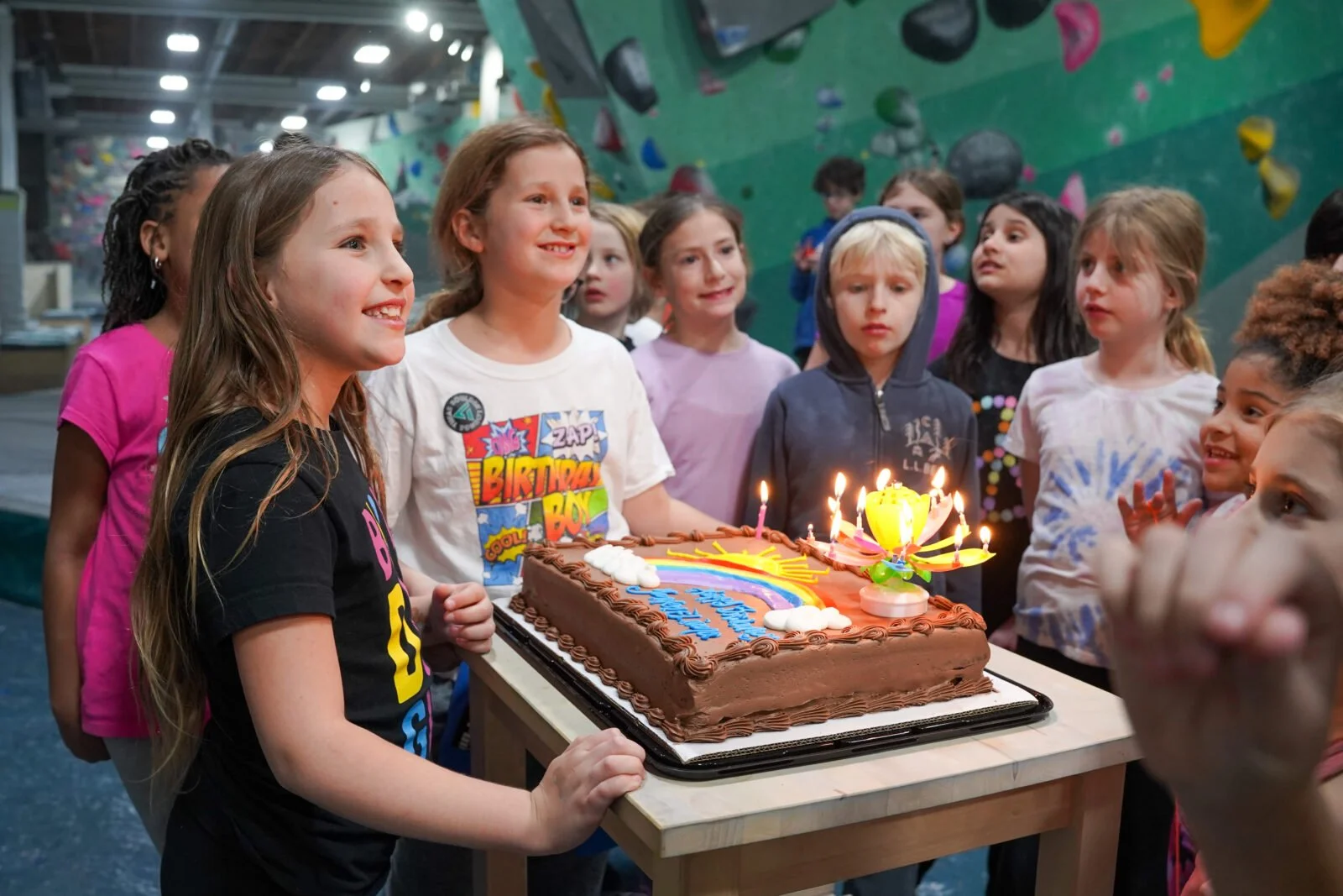 Children gathered around a birthday cake with lit candles at a birthday celebration in an indoor sports or climbing facility.