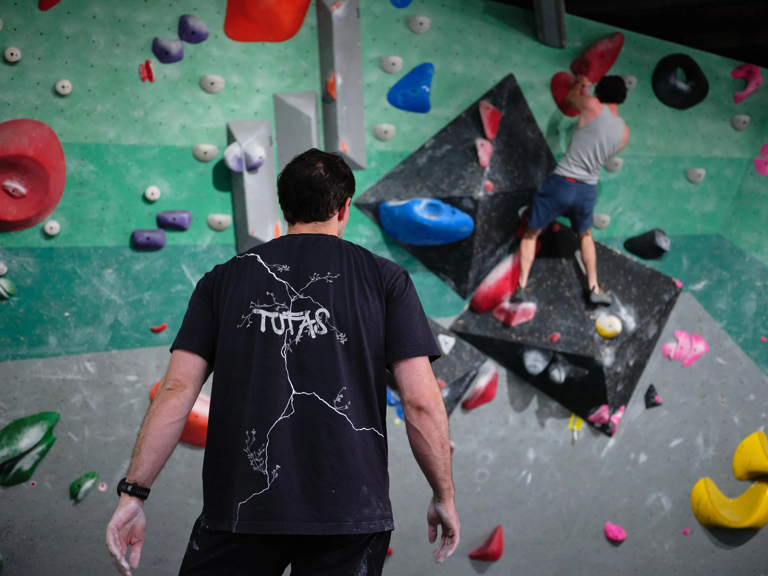 Indoor bouldering gym with colorful climbing holds, a climber in a tank top and shorts, and a person observing in the foreground, wearing a black T-shirt with white text and lightning bolt design.