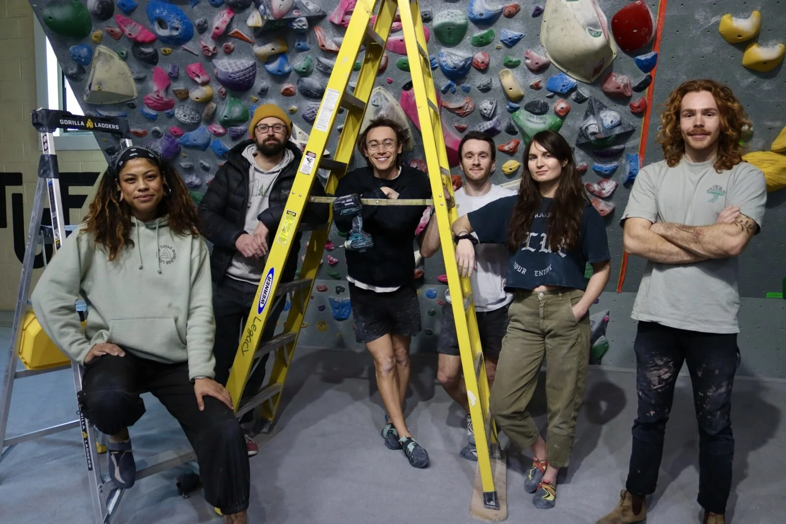 Group of seven people at an indoor rock climbing gym, standing in front of a colorful climbing wall. They are posing with a ladder and a cordless drill, suggesting they are working on or preparing for climbing wall maintenance or setup.