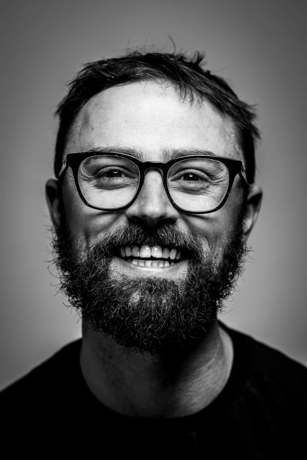 Black and white portrait of a smiling man with glasses and a beard, wearing a dark shirt.