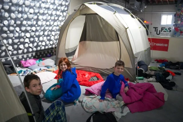 Three children sitting on sleeping bags in front of a large camping tent indoors, with climbing wall and sports banners in the background.