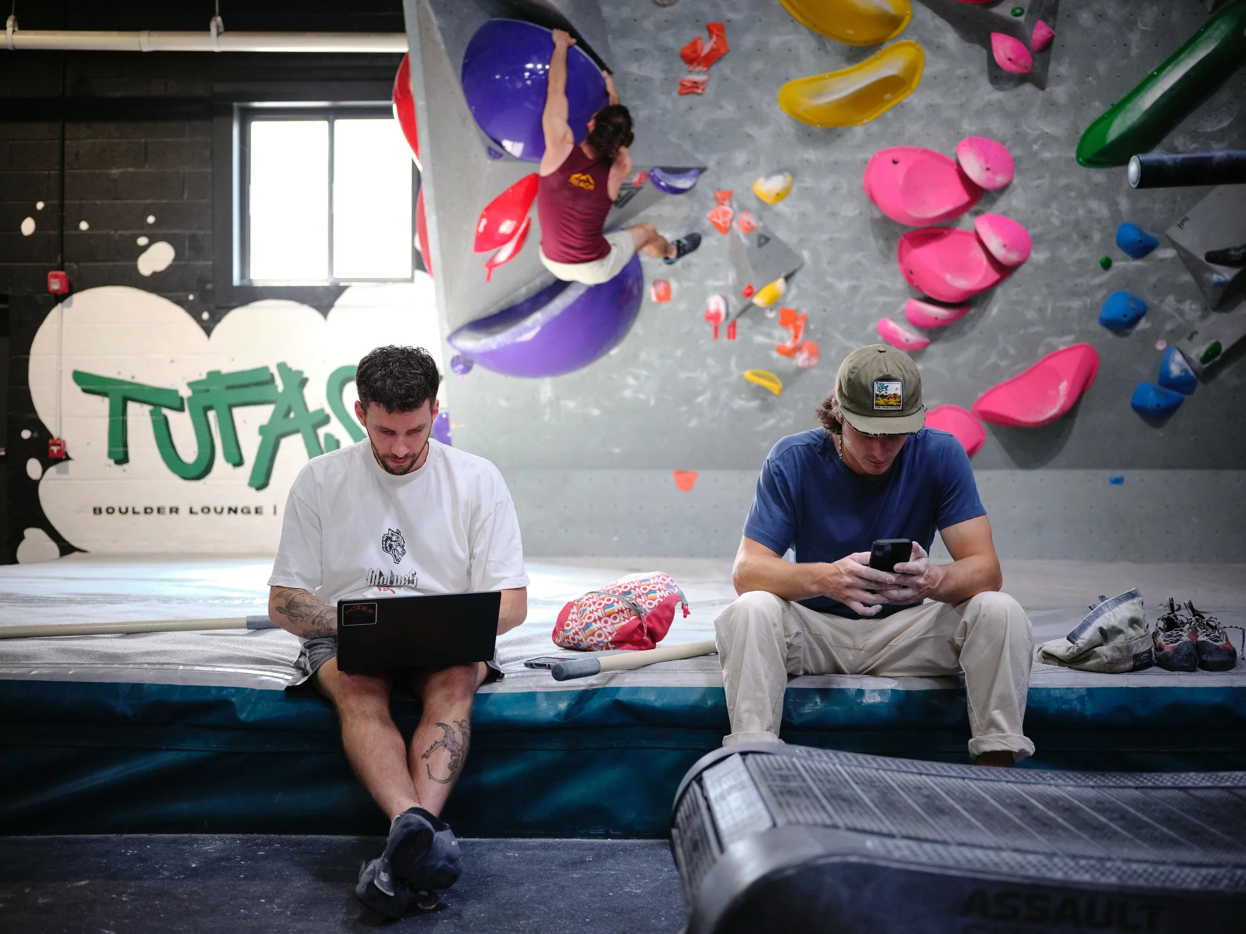 Two men sitting on the floor in a climbing gym, looking at their phones, with a climber on a wall behind them. The climber is reaching for holds on an indoor bouldering wall with colorful holds.