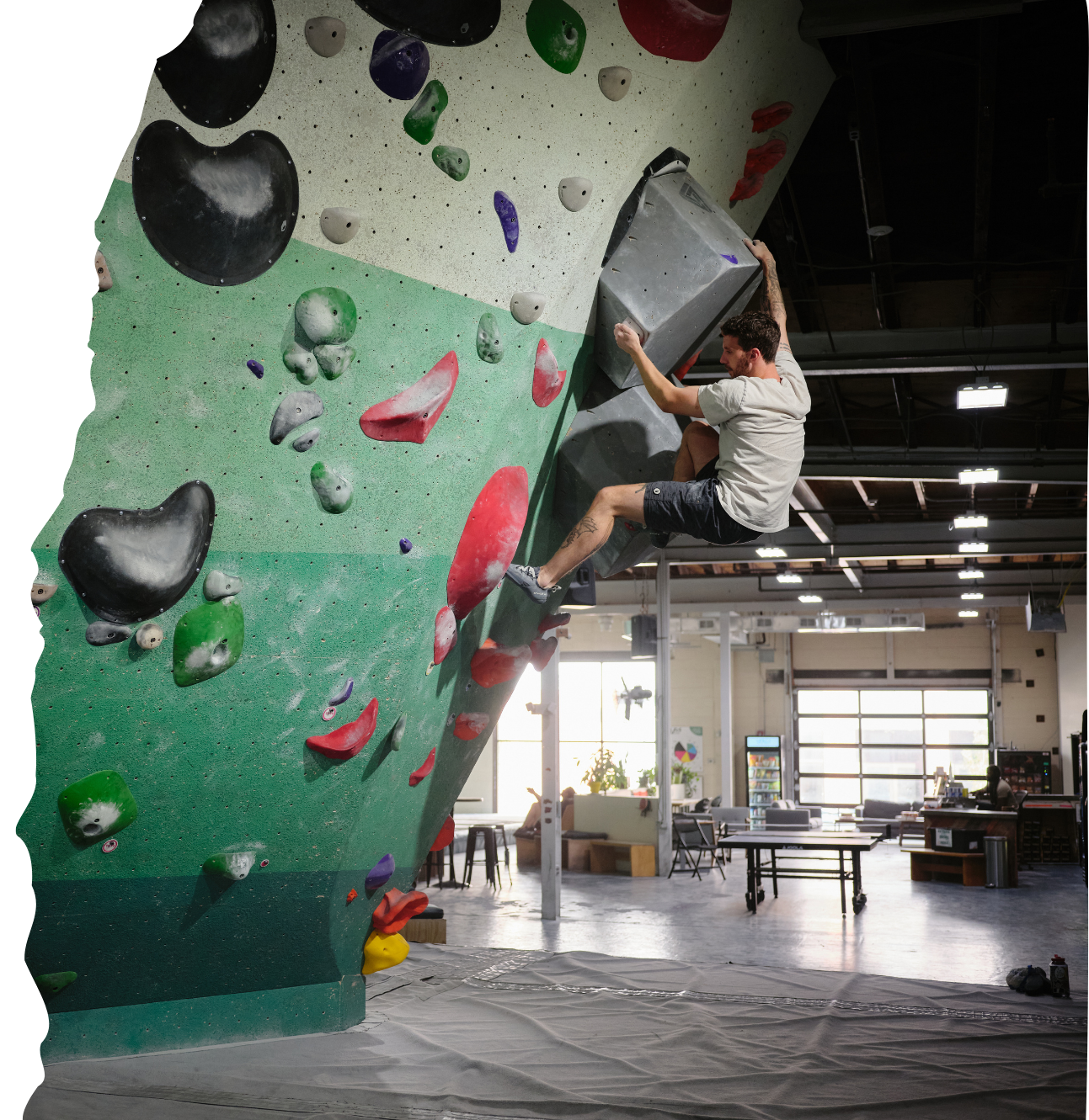 An indoor bouldering gym with a man climbing on a tilted green climbing wall with colorful holds, sitting on a large gray volume.
