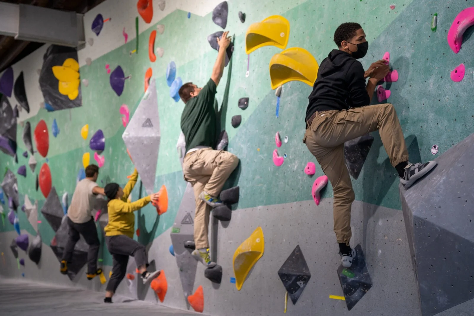 Four people, wearing face masks, climbing on an indoor bouldering wall with colorful holds in various shapes.