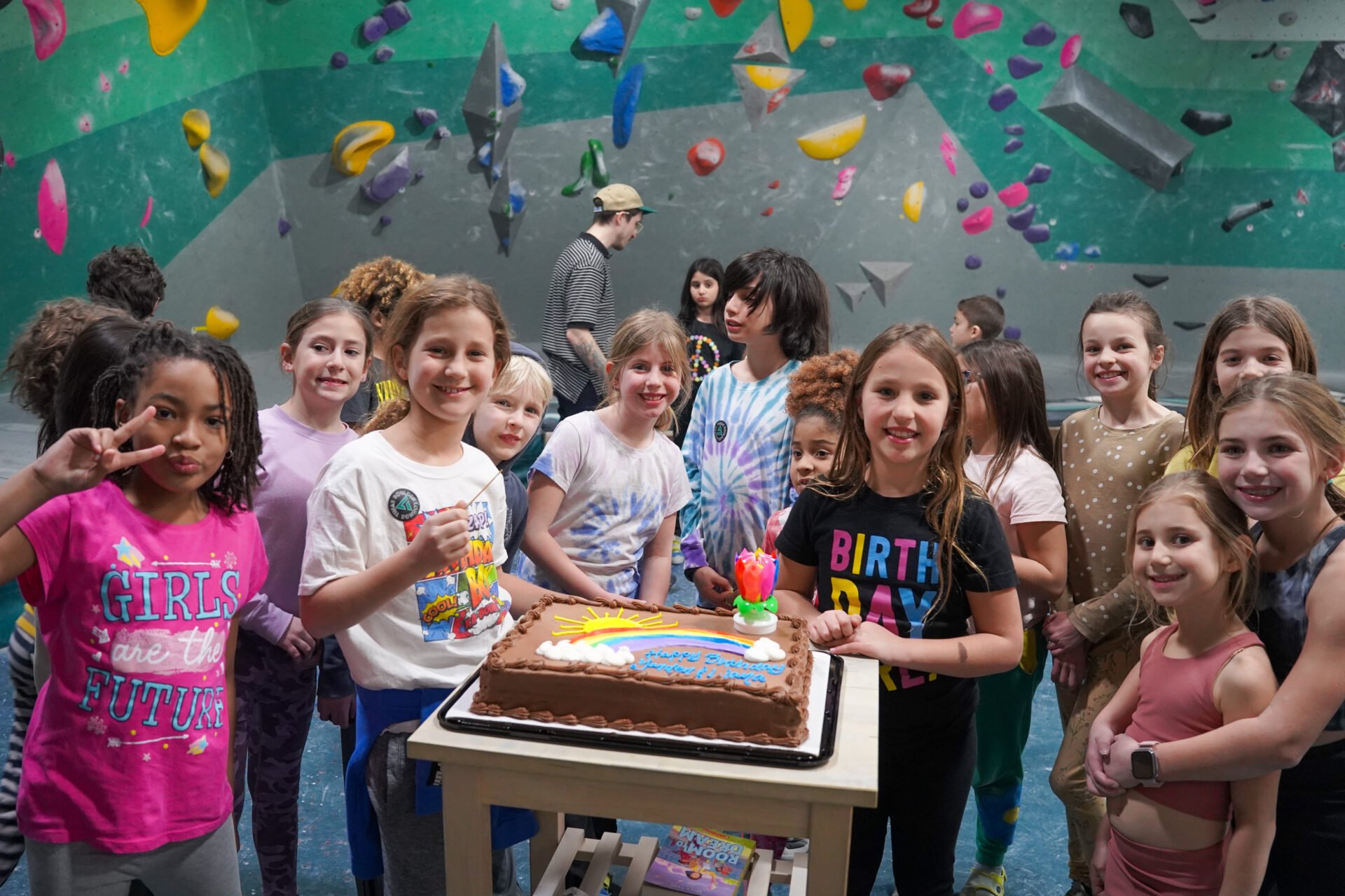 A group of children celebrating a birthday party in an indoor climbing gym, gathered around a table with a large birthday cake decorated with a rainbow, clouds, and a sun. The children are smiling and wearing colorful clothes.