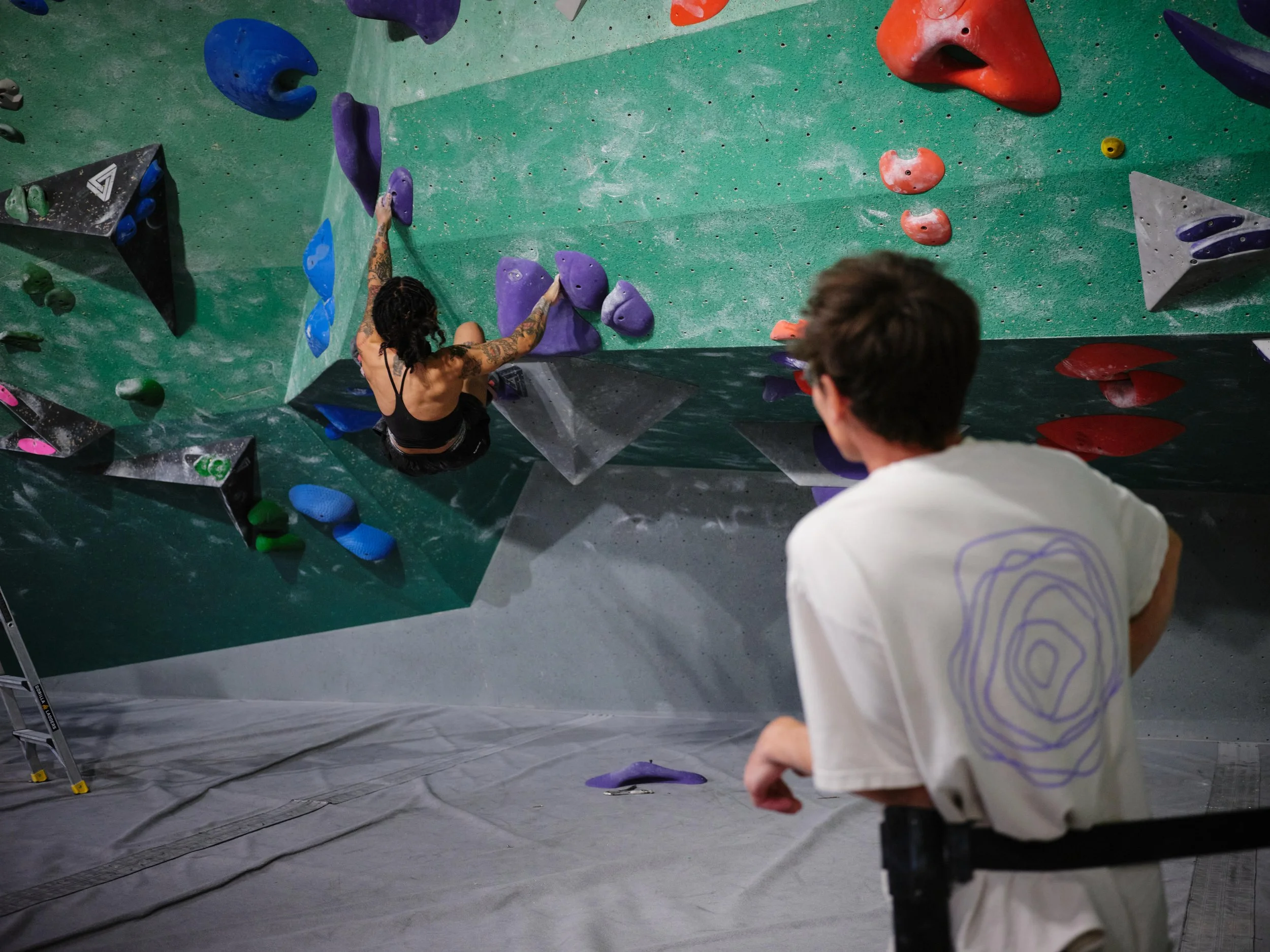 A woman with tattoos climbing an indoor bouldering wall with colorful holds, supervised by a man in a white shirt.