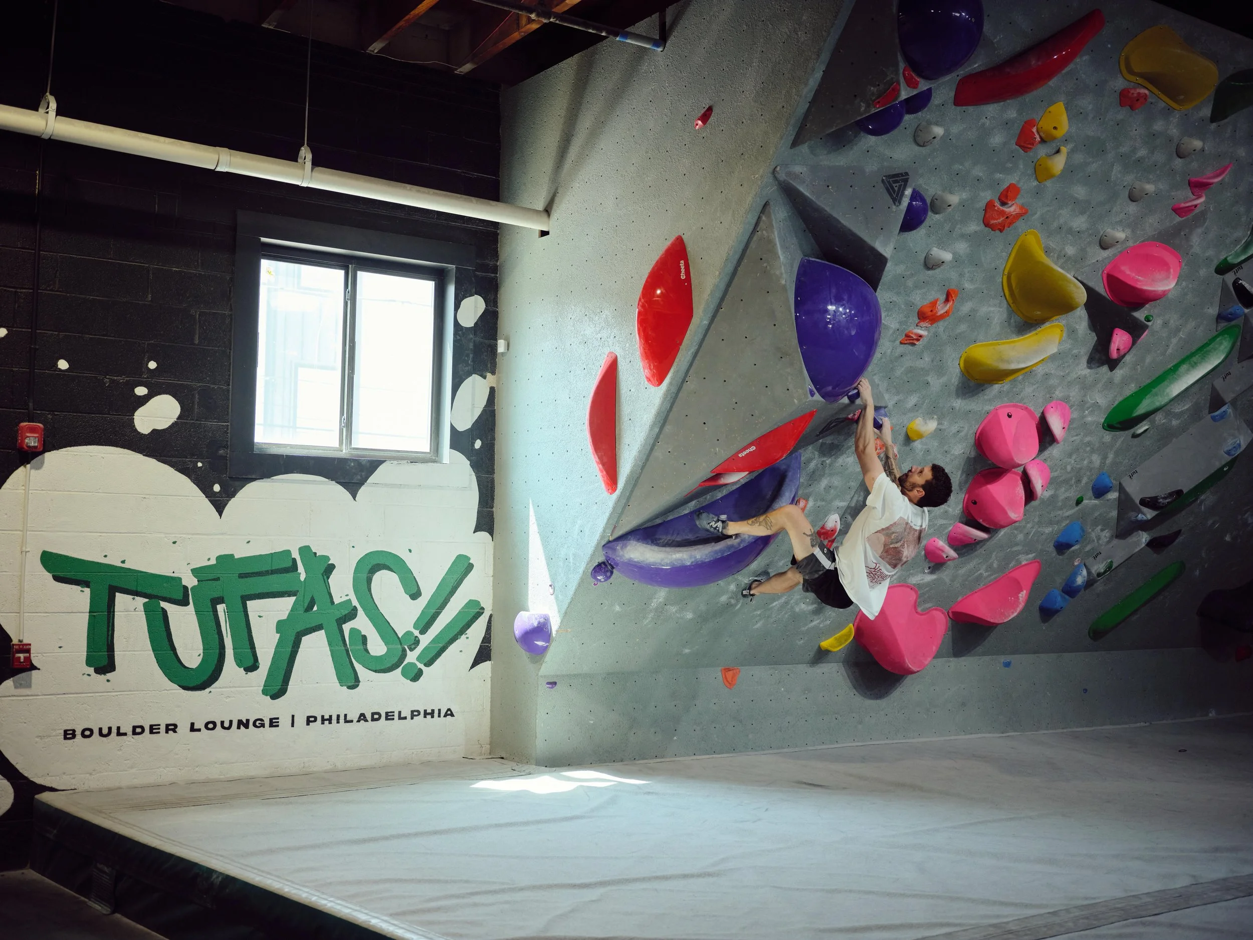 Indoor climbing gym wall with various colorful holds. A person is lying on the floor near the wall, wearing a white shirt and black shorts, with a tattoo on their right calf. The wall has a sign that says 'TUTS!' and mentions 'Boulder Lounge Philadelphia'.
