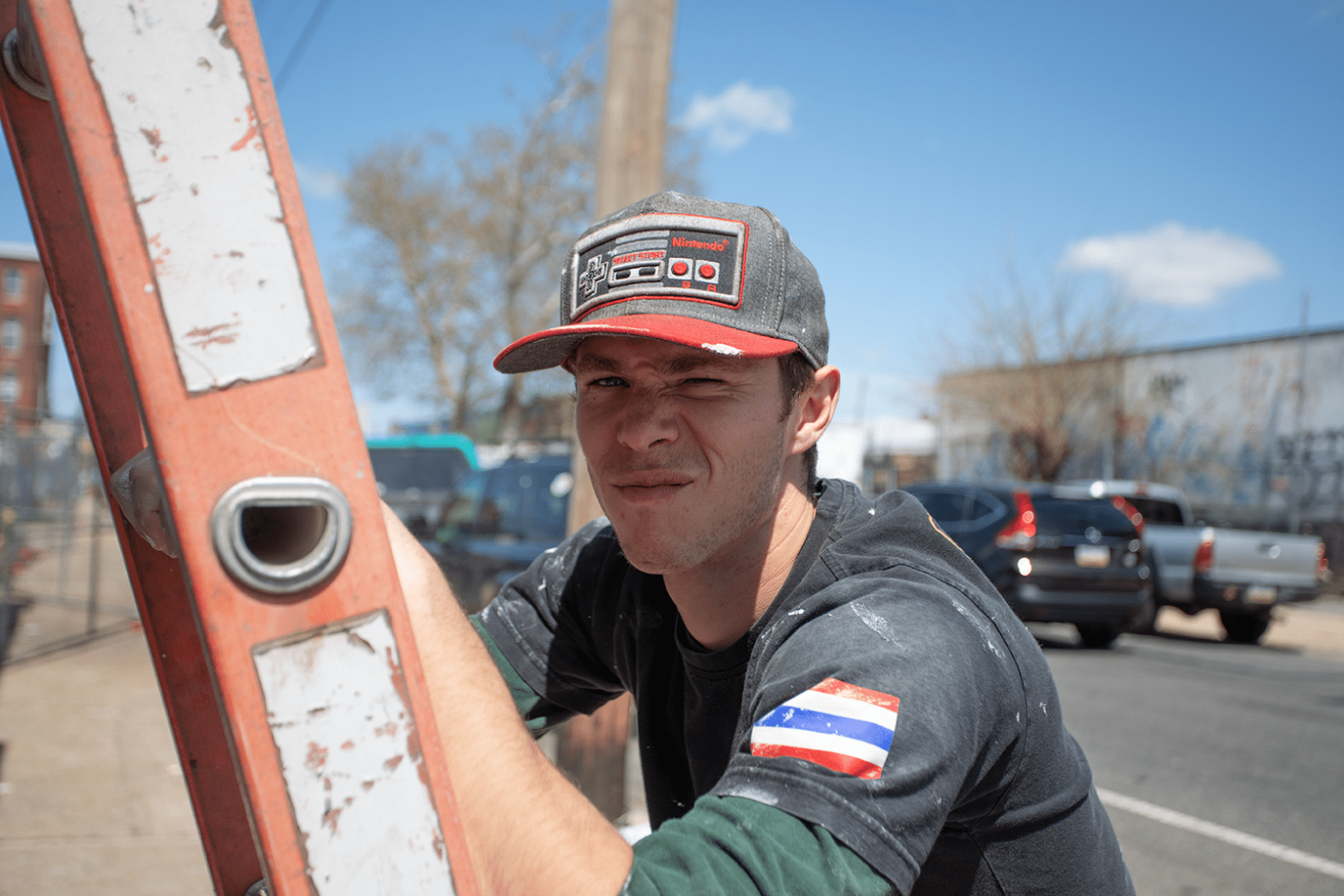 A young man in a black shirt with a Costa Rican flag patch on the sleeve, wearing a gray and red Nintendo cap, is leaning on an orange ladder with a serious or skeptical expression. Vehicles and trees are visible in the background on a sunny day.