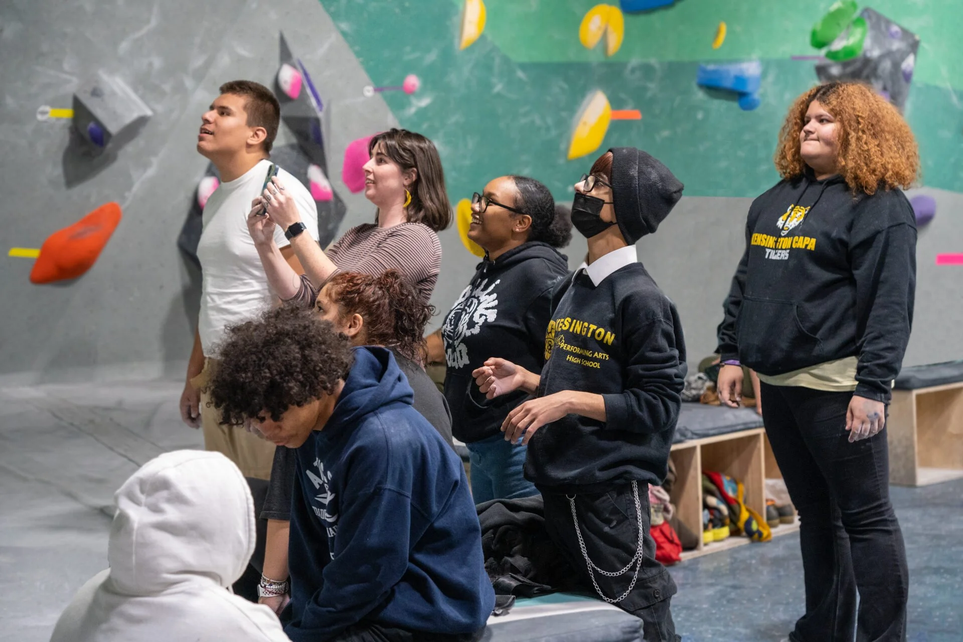 A group of young people, some standing and some kneeling in a climbing gym, with colorful climbing holds on the wall behind them. Some are wearing hoodies or masks.