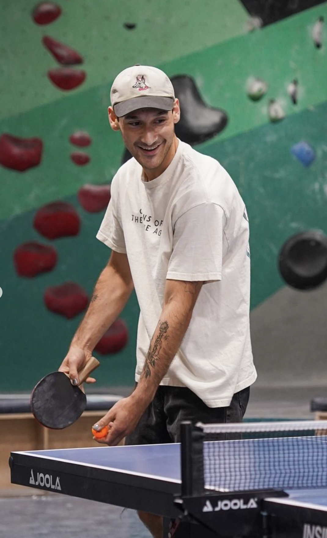 A young man with tattoos on his arms playing table tennis in a sports facility with a green rock climbing wall in the background.