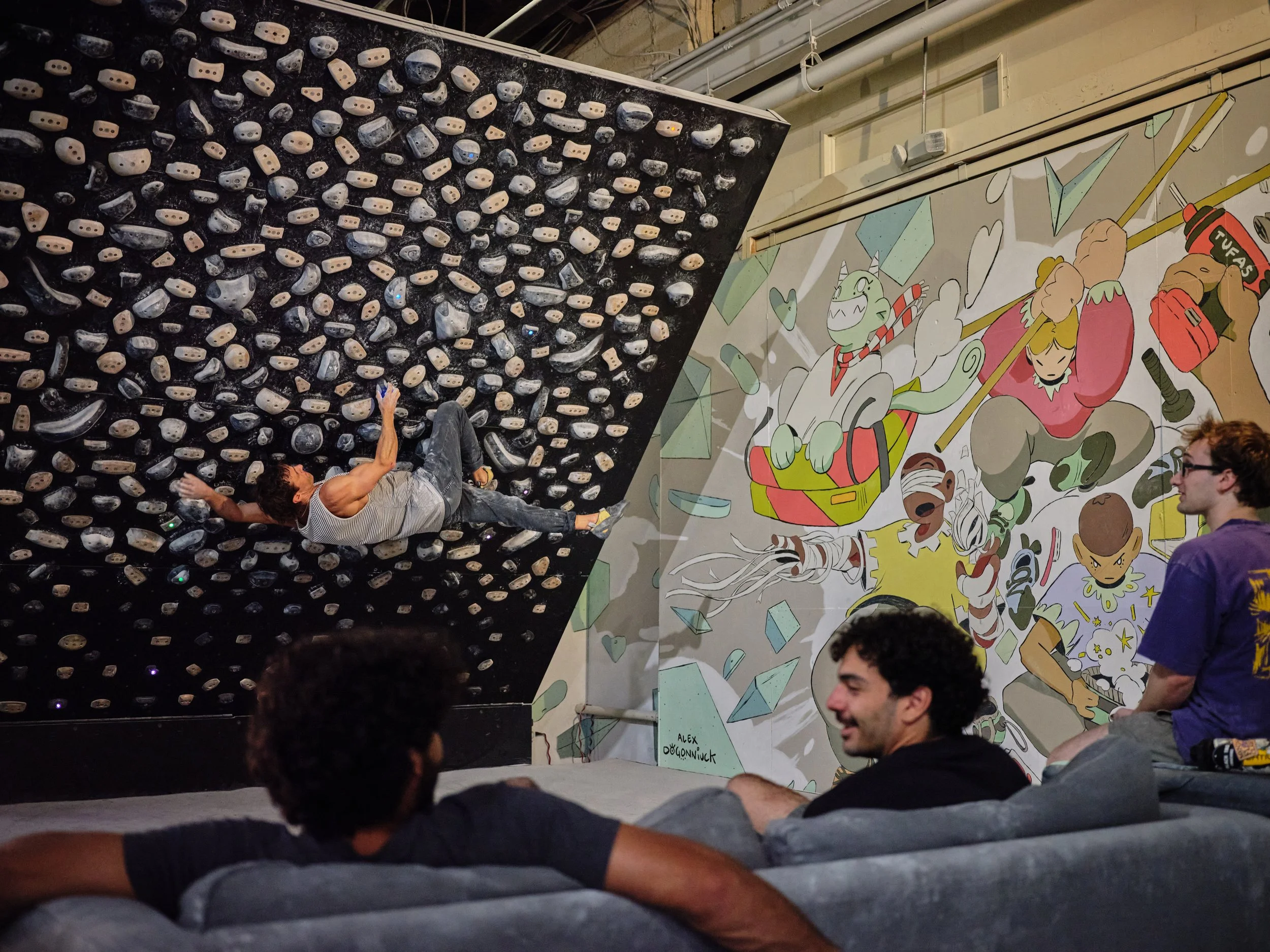Indoor rock climbing gym with a man reaching for climbing holds on a black bouldering wall, and several people watching. The wall has numerous beige, black, and gray holds, and there is a colorful mural on the adjacent wall depicting cartoon characters and geometric shapes.