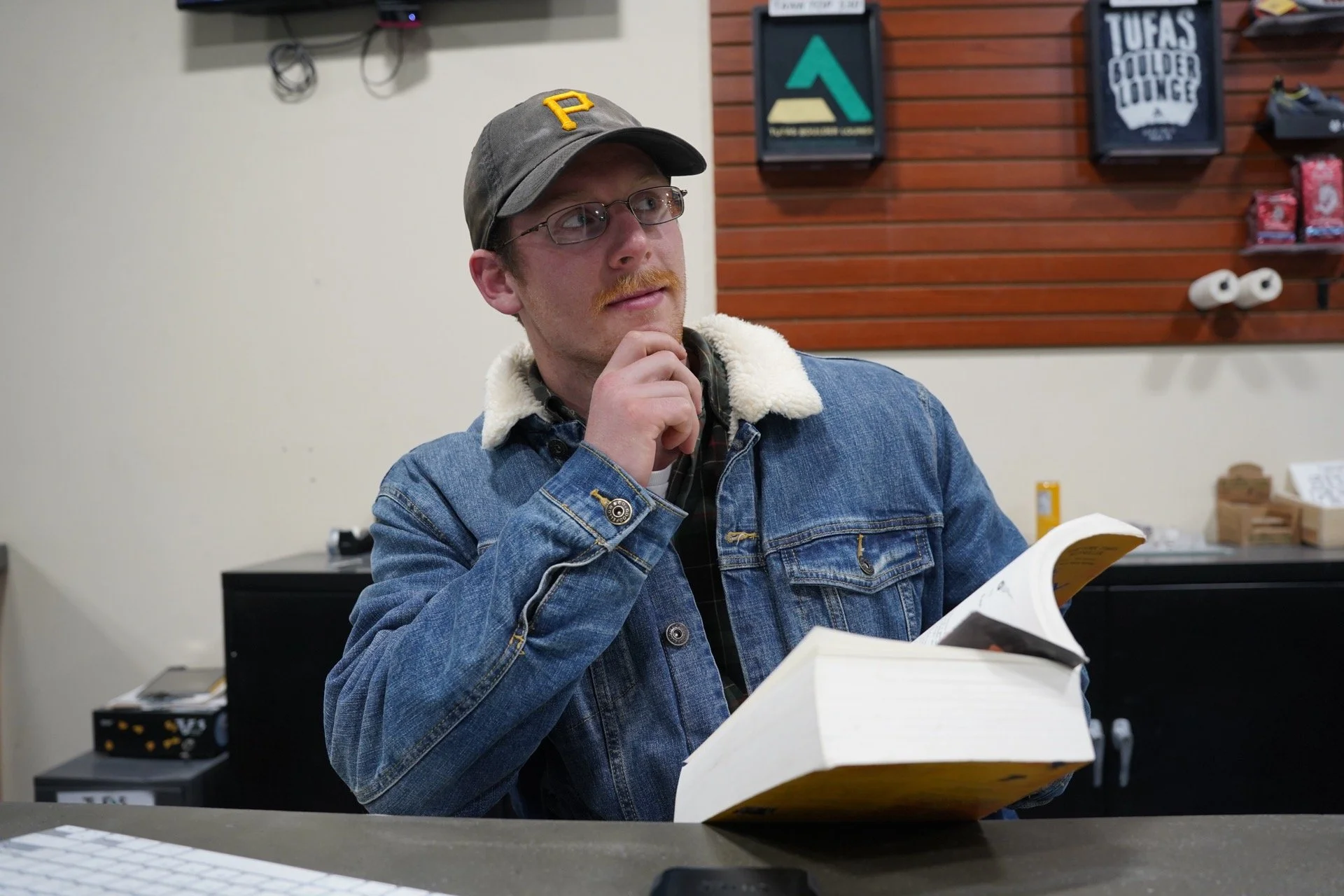 A young man with glasses and a mustache, wearing a gray baseball cap with a yellow 'P' and a denim jacket with a sherpa collar, is sitting at a table in a retail or cafe setting, holding an open book or menu and thinking. In the background, there are shelves with packaged products, a framed sign reading 'TUFA'S COLDER LOUNGE,' and electronic devices mounted on the wall.