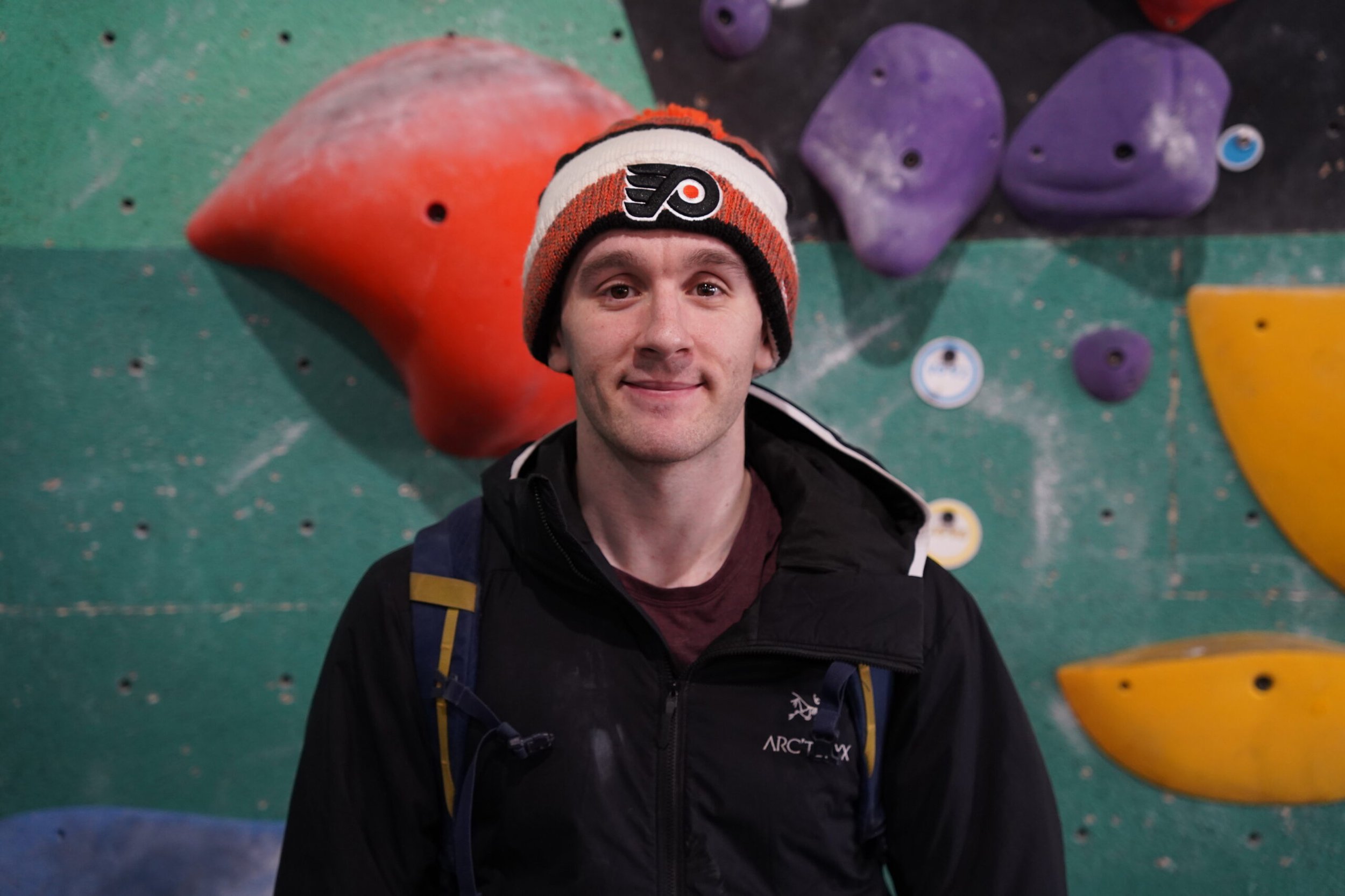 Young man smiling, wearing a black jacket, and a colorful knit hat with a Philadelphia Flyers logo, standing in front of a bouldering wall with multicolored climbing holds.