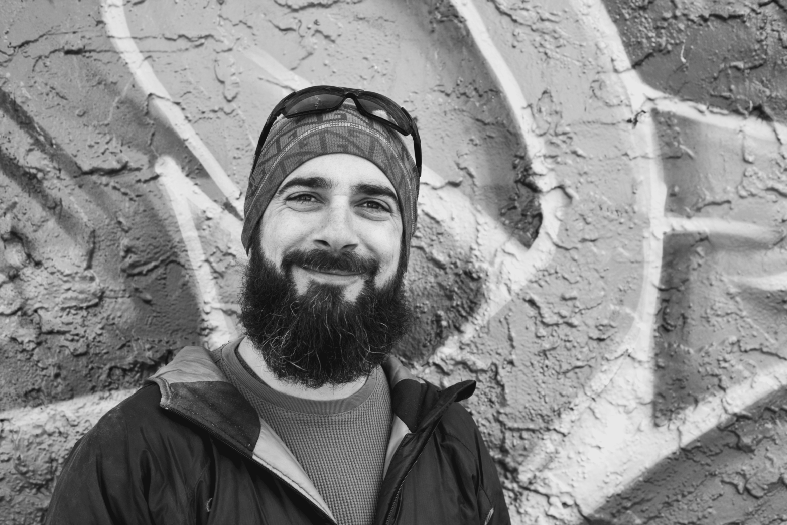 A smiling man with a beard and mustache wearing a beanie, sunglasses on top and a jacket, standing in front of a wall with graffiti.
