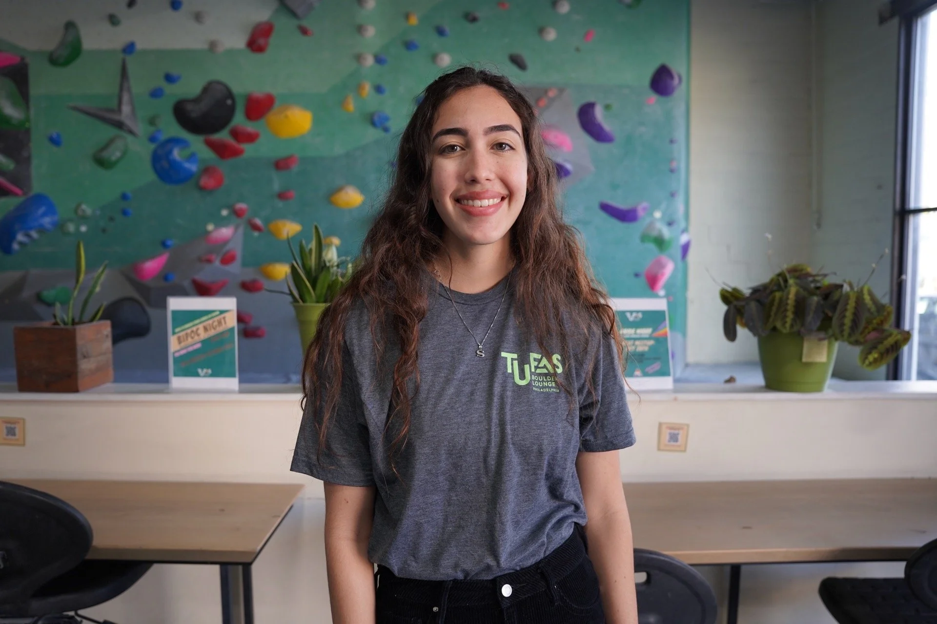 Young woman with long, curly brown hair smiling, wearing a dark gray T-shirt with a logo, standing in a climbing gym with colorful holds on the wall behind her and potted plants on a counter.